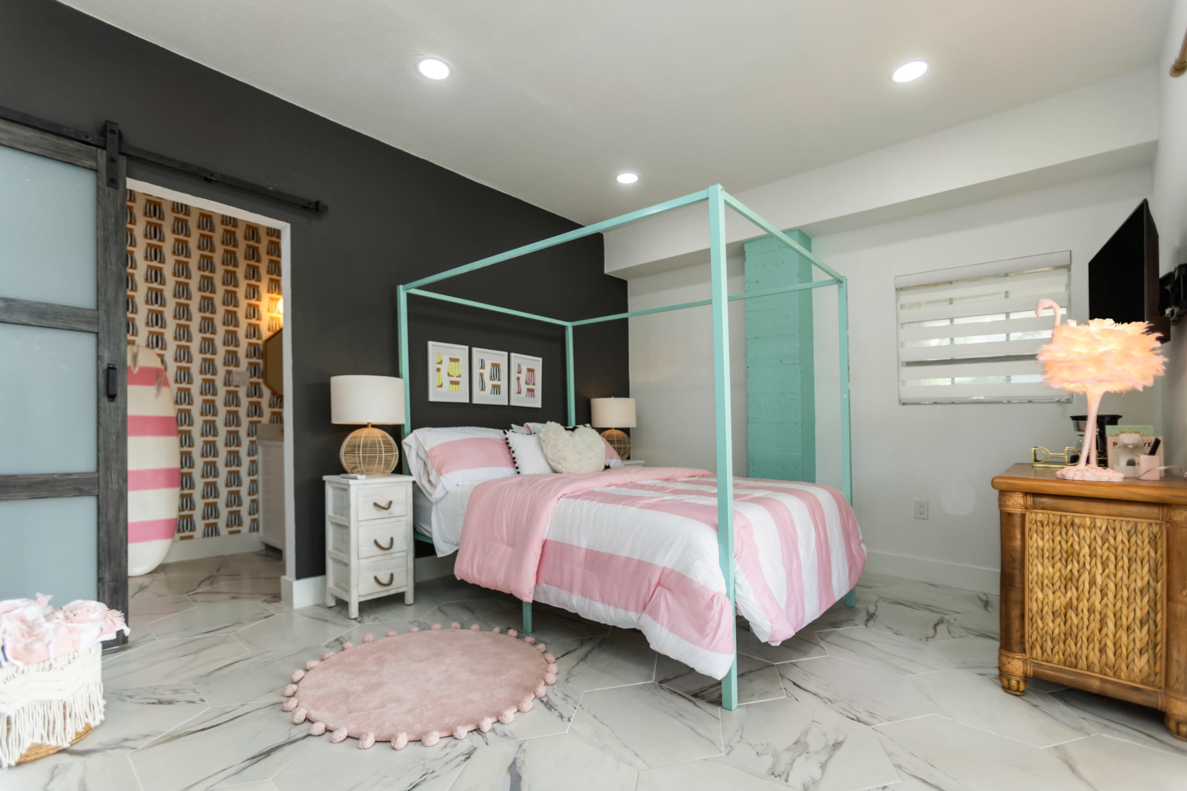 The image shows a modern bedroom with a teal four-poster bed, pink and white bedding, and light-colored furnishings, including a nightstand and a decorative rug.