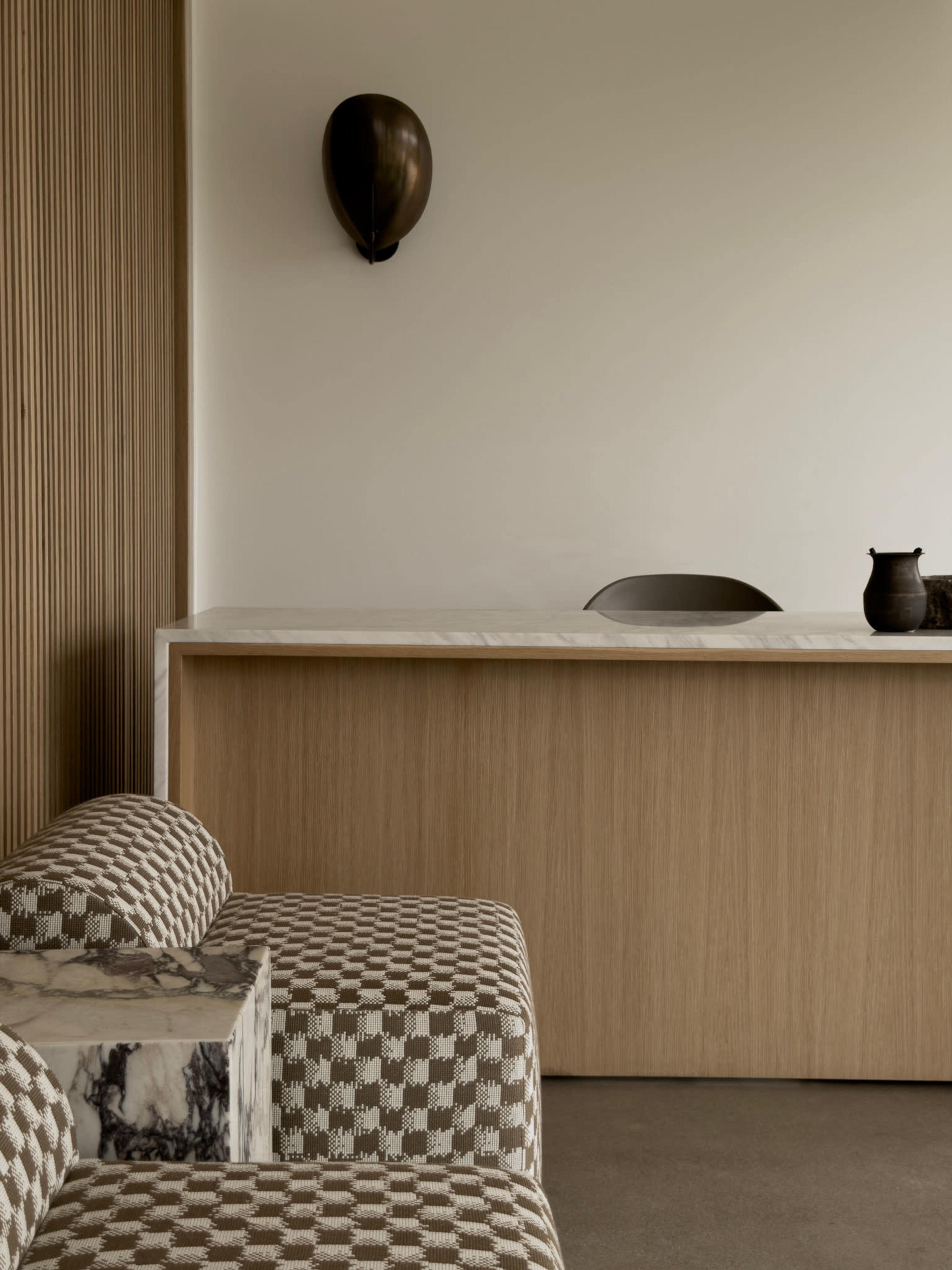 The image shows a minimalist interior with a wooden reception desk, a two-tone patterned couch, and a wall-mounted decorative piece.