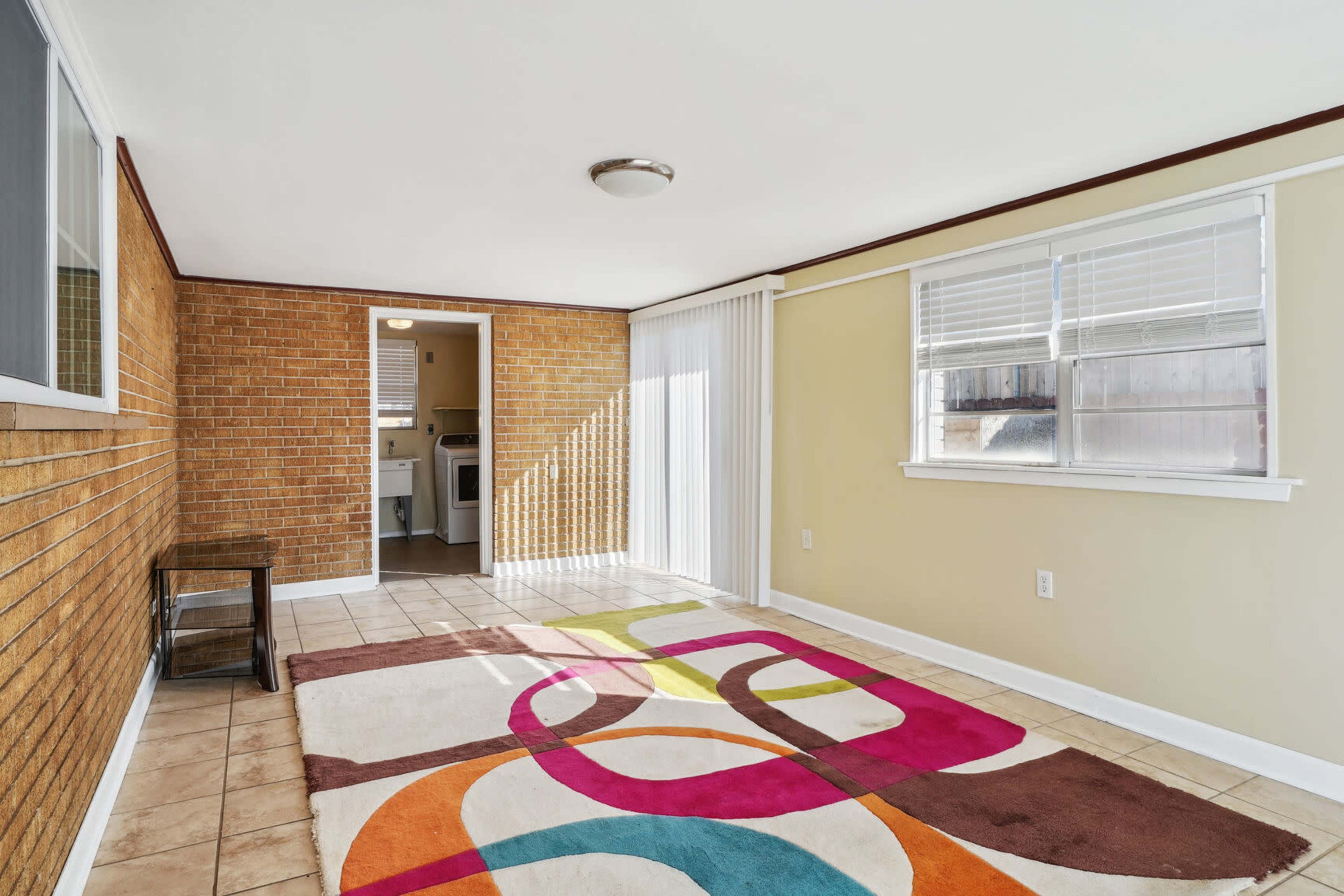 The image shows a sunlit room with brick and painted walls, featuring a colorful area rug and a doorway leading to a laundry area.