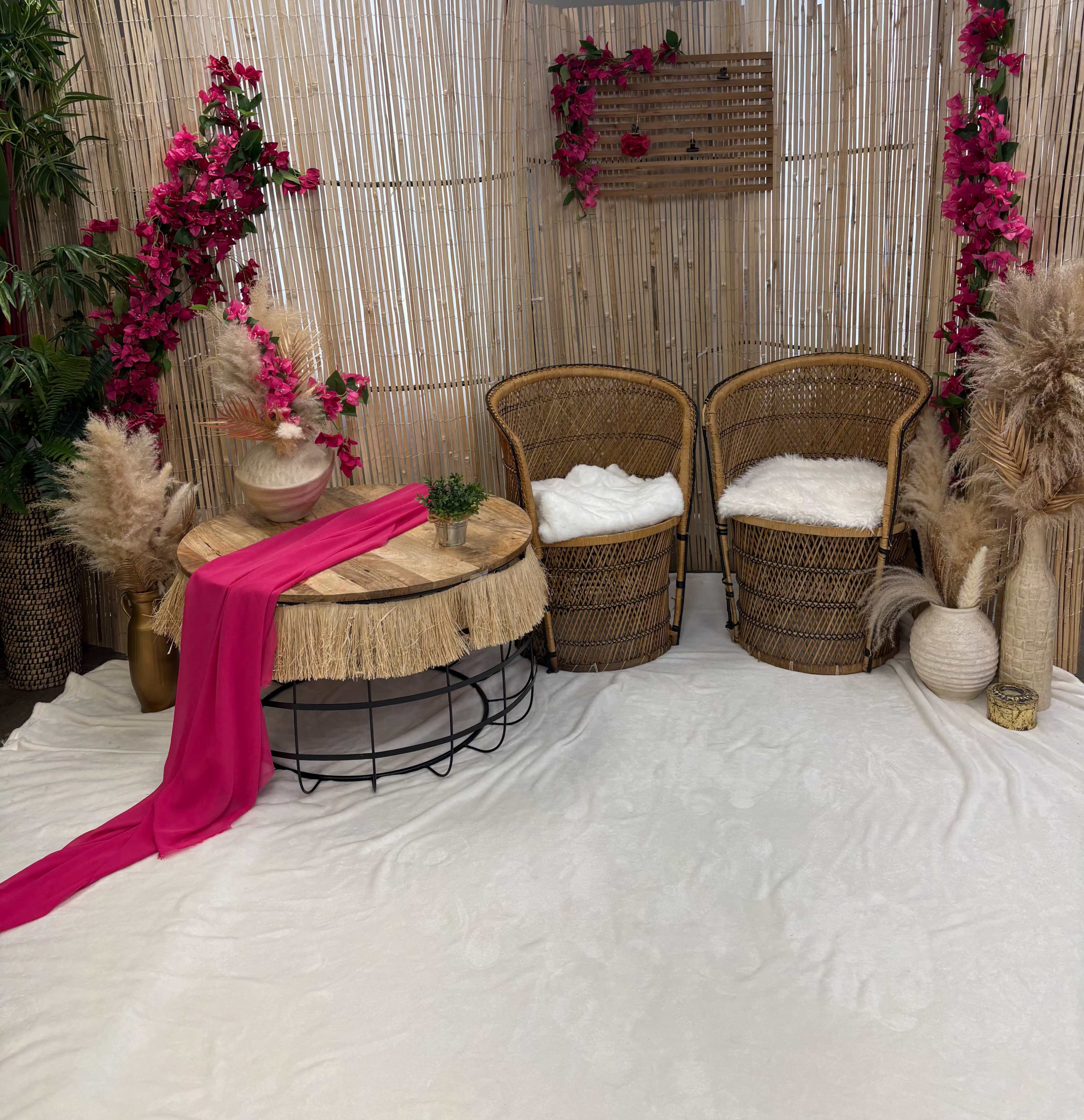 The scene features two wicker chairs, a round table with a fringed edge, and decorative plants arranged against a bamboo backdrop adorned with pink flowers.
