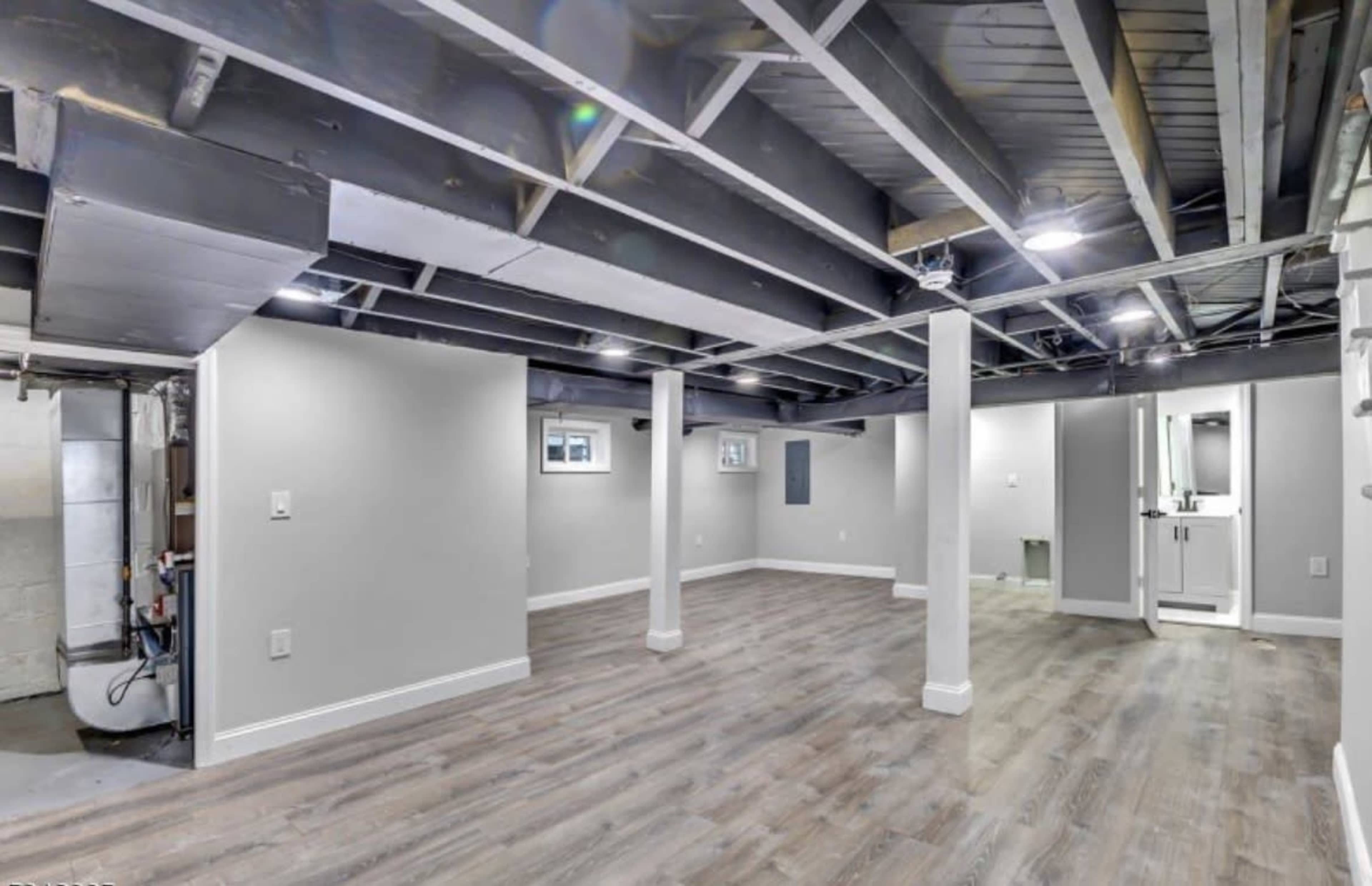The image shows a vacant basement with wooden flooring, exposed beams, and light gray walls.