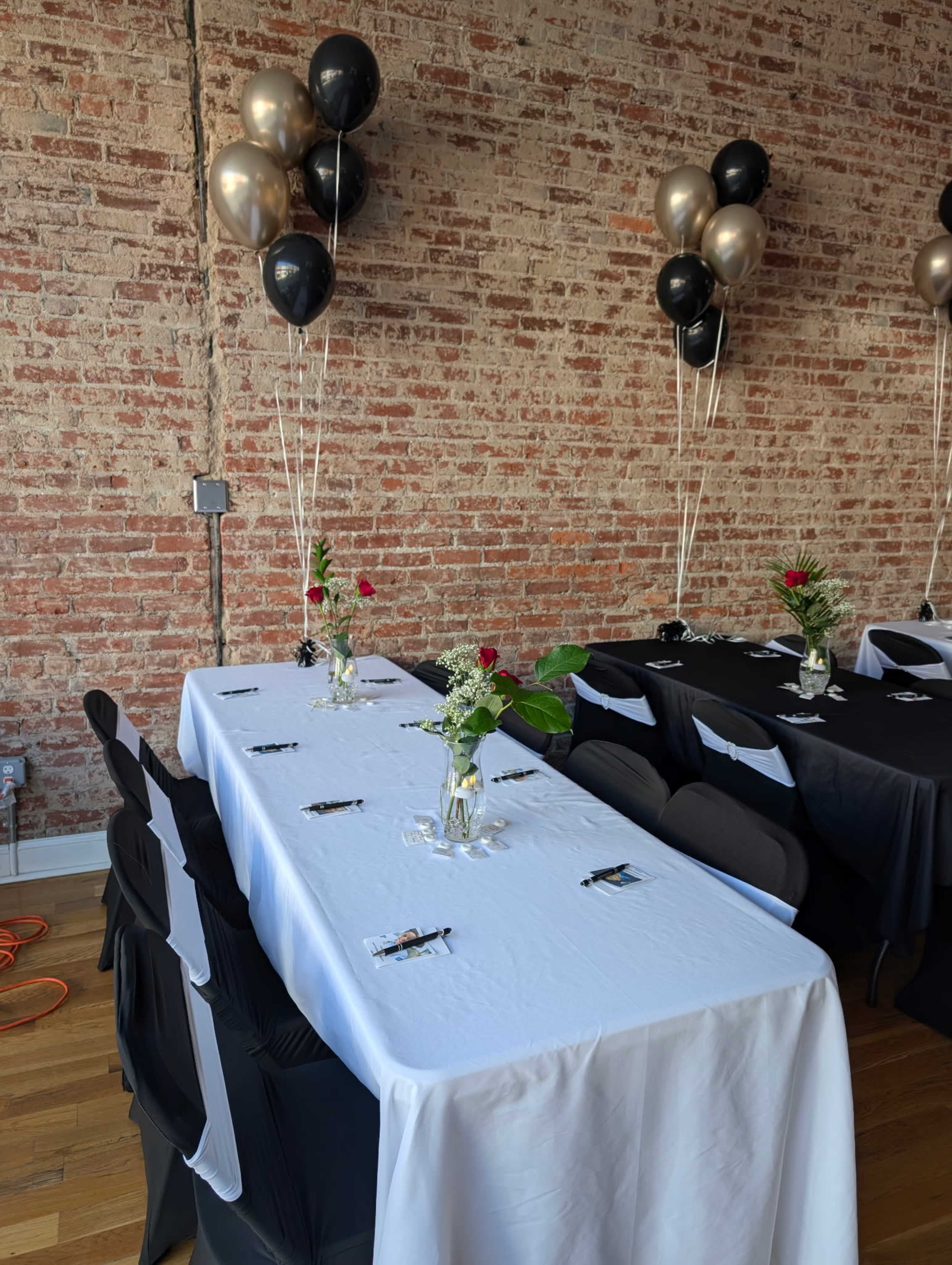 A long table is set with white and black tablecloths, adorned with small vases of flowers and balloons in black and gold, against a brick wall.