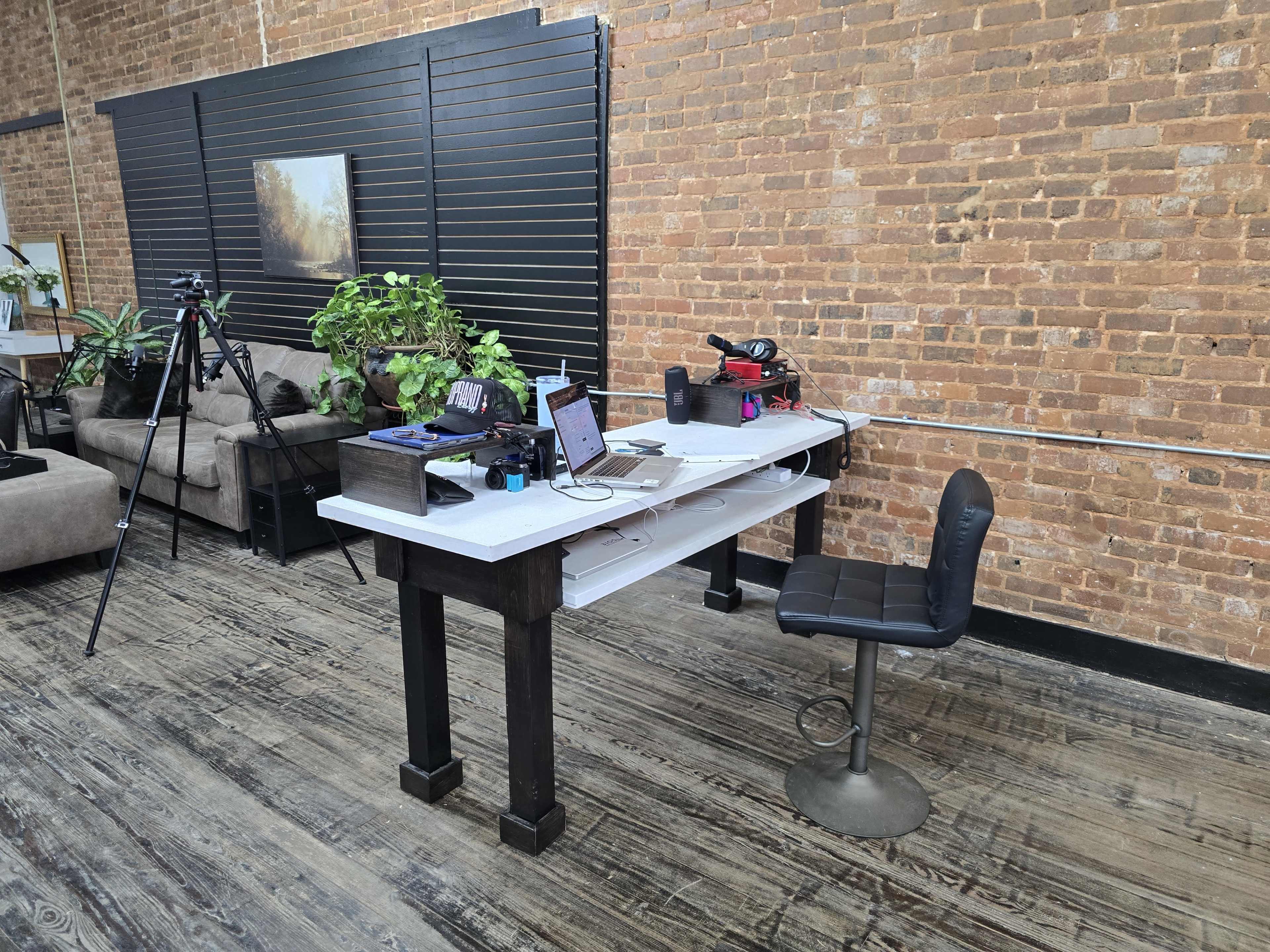 A workspace features a white desk with electronic equipment, a chair, and potted plants in a room with exposed brick walls.