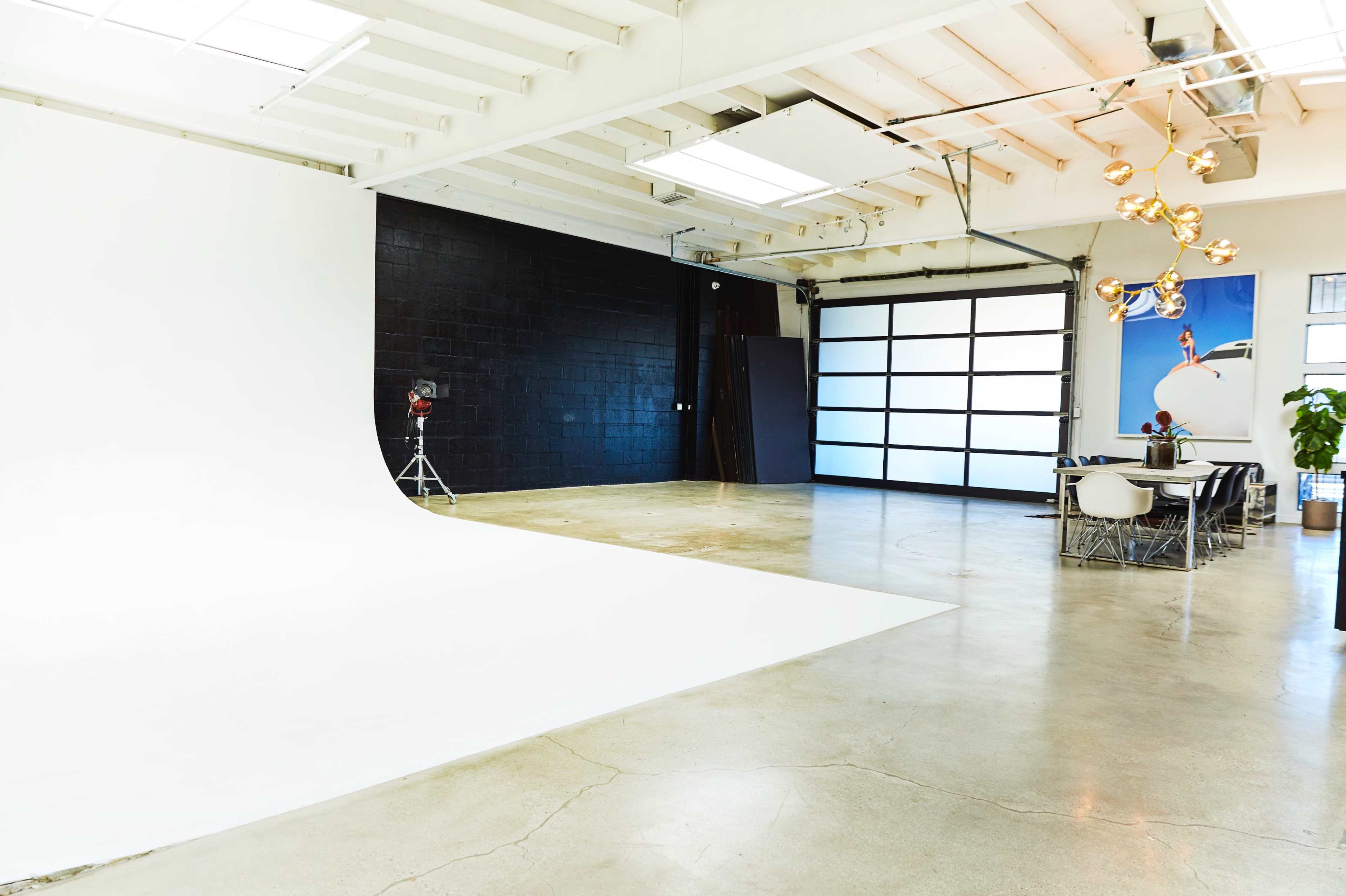 The image shows a spacious photo studio with a white backdrop on one side and a modern dining area with a large table and decorative lighting on the other.