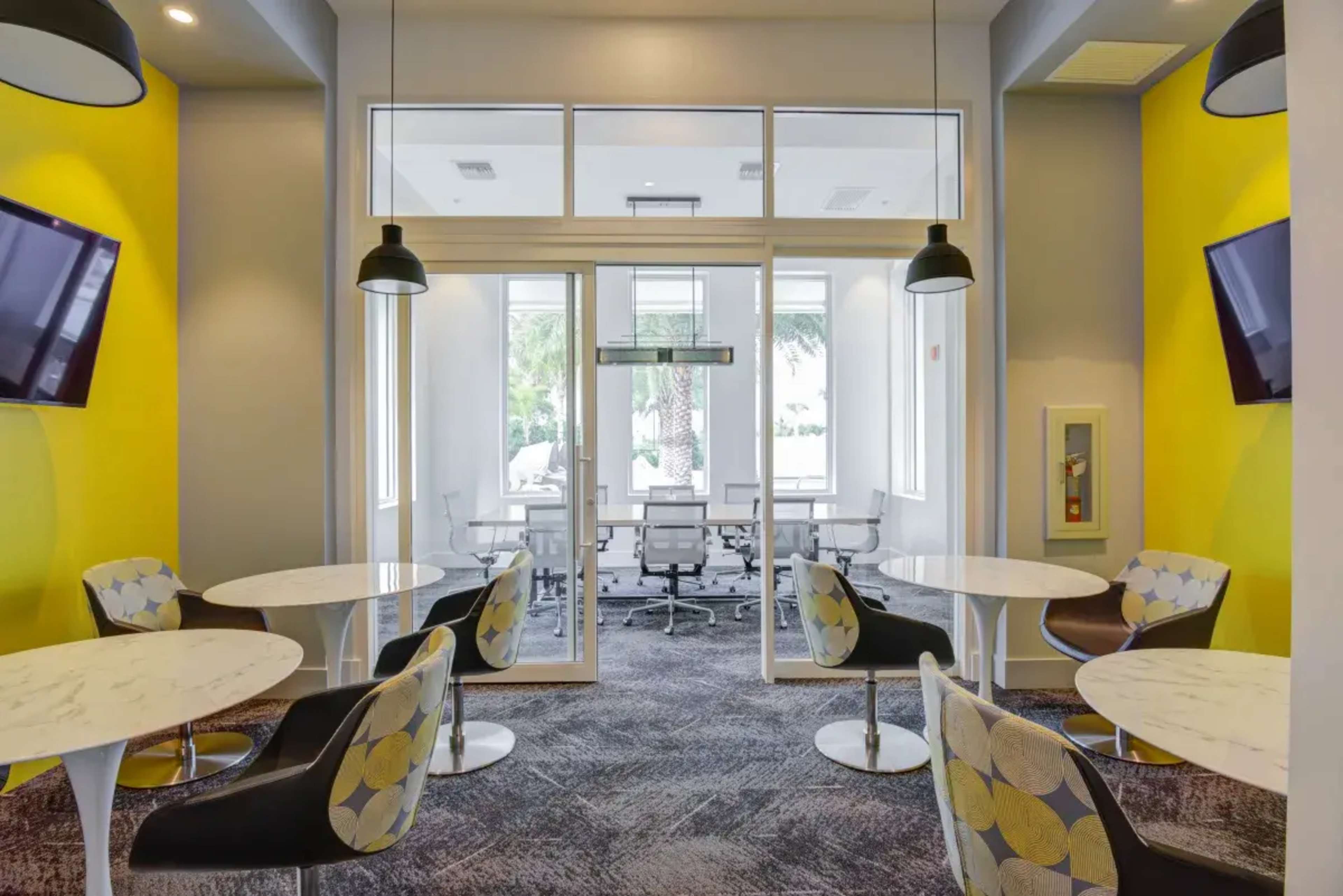 The image shows a modern office space featuring round tables, chairs, and two large screens on vibrant yellow walls, with glass doors leading to a meeting room.