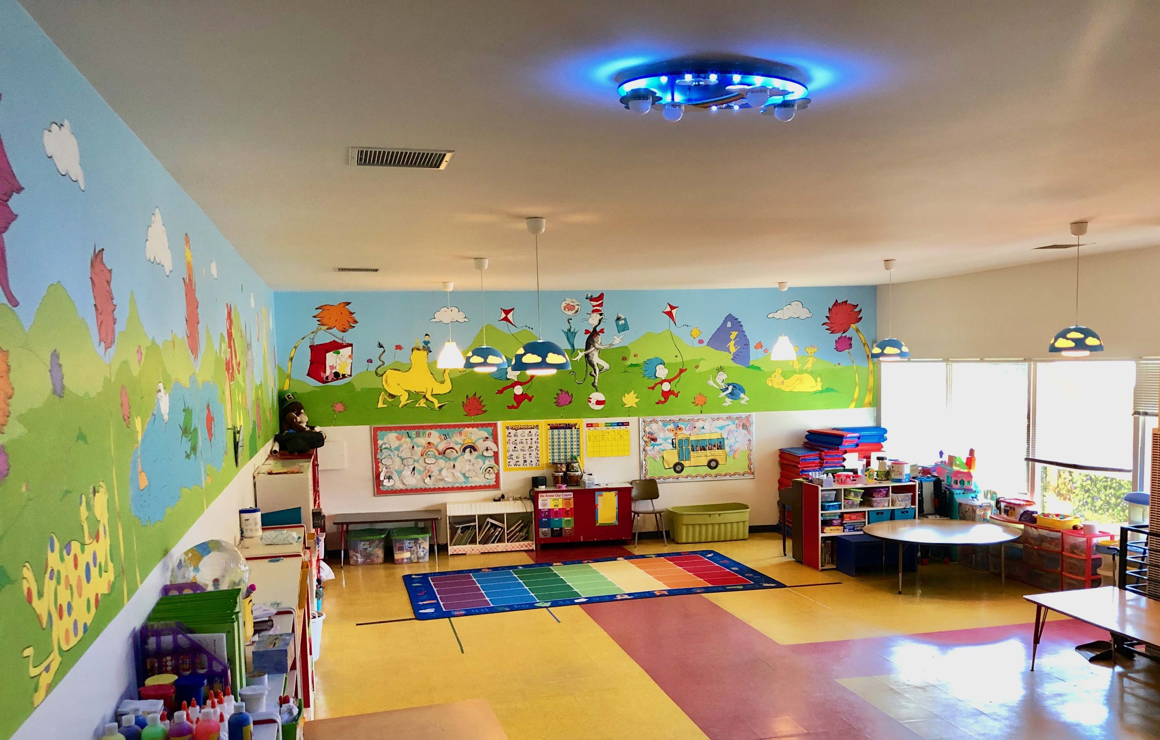 A brightly colored classroom features a mural of animals and landscapes, with tables and storage areas for educational materials.