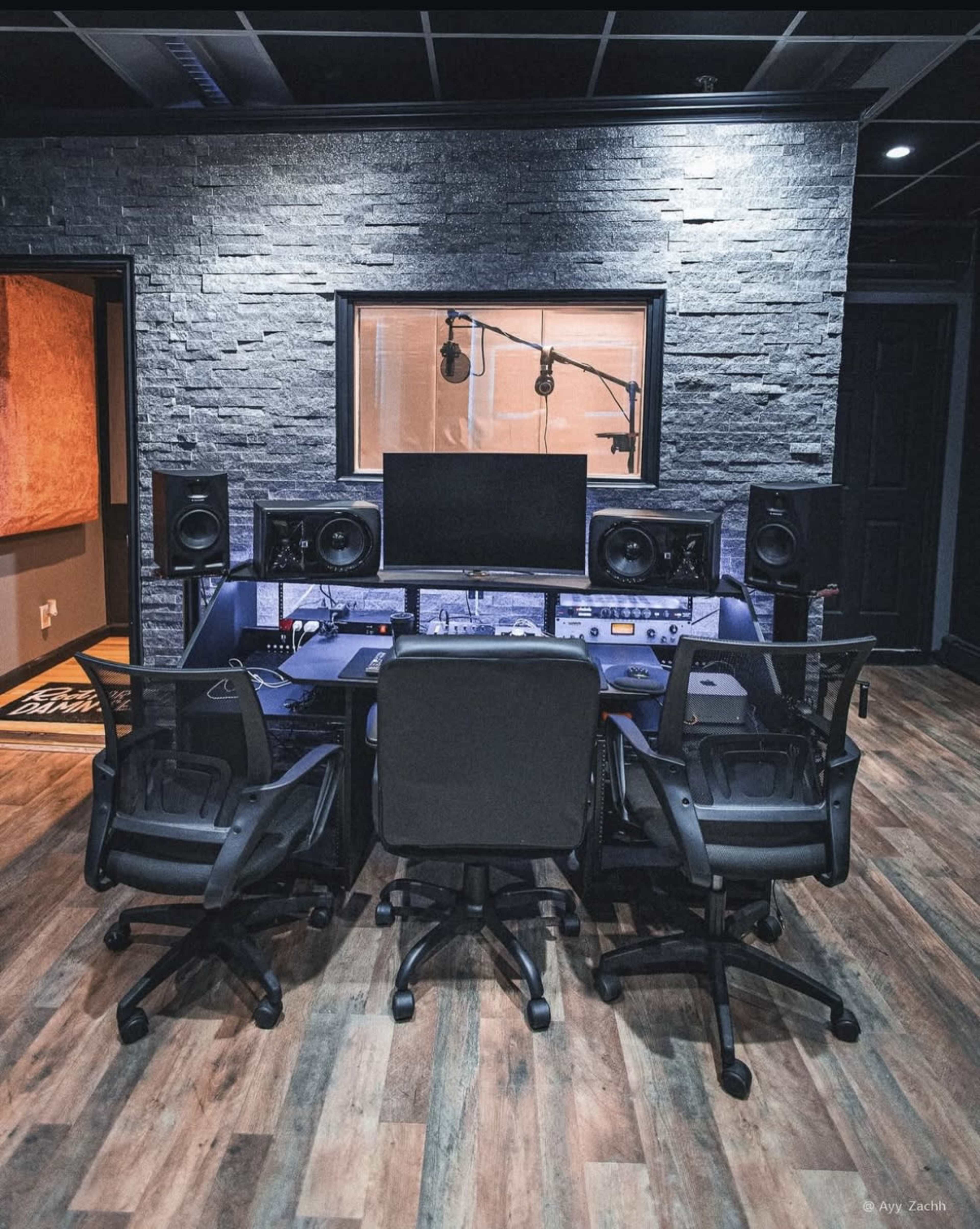 The image shows a modern music studio featuring a central mixing desk with three chairs, audio monitors, and a glass window overlooking a recording area.