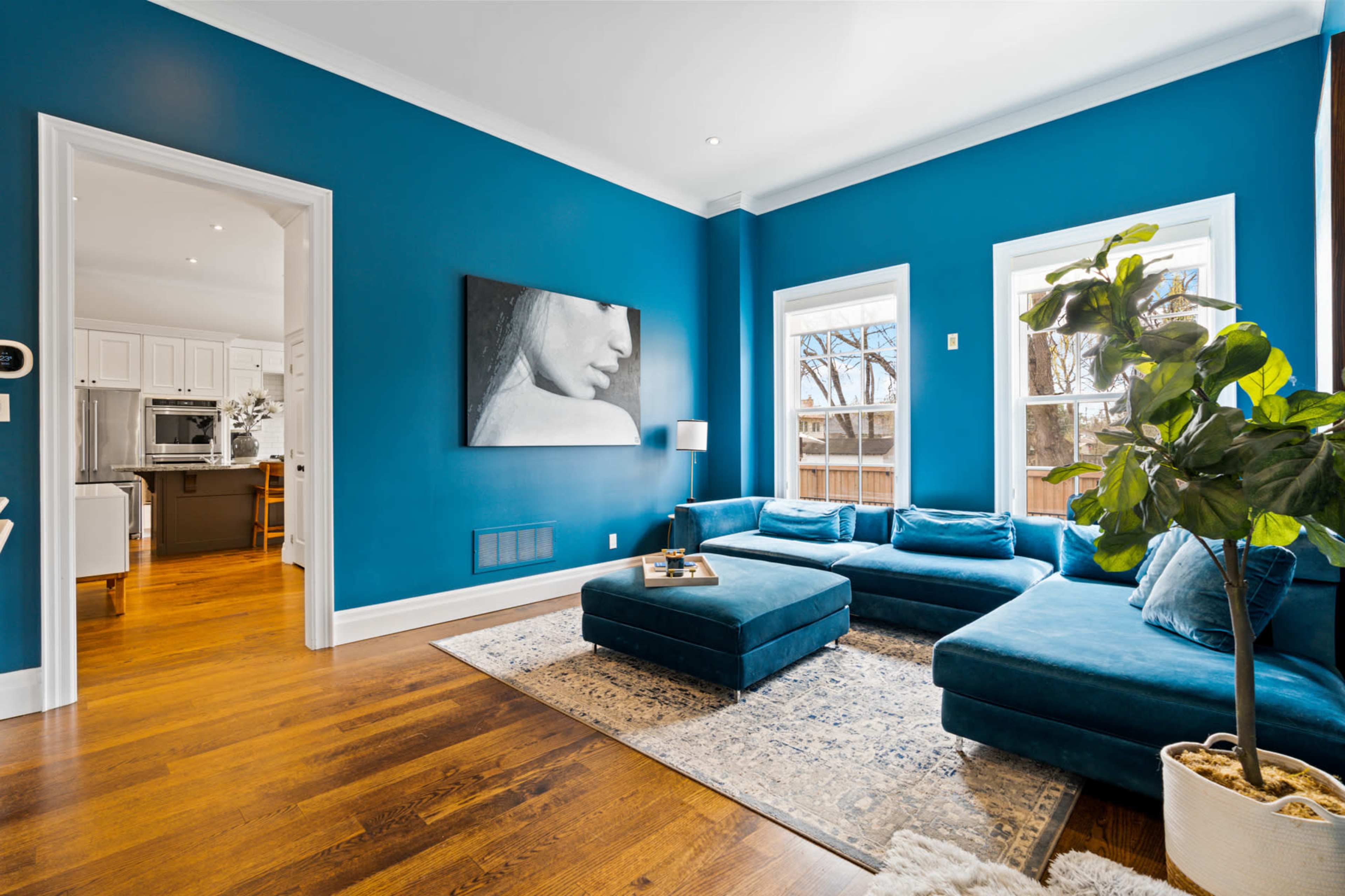 A living room with a blue accent wall, a large sectional sofa, and a decorative area rug, leading to an adjacent space with a kitchen.