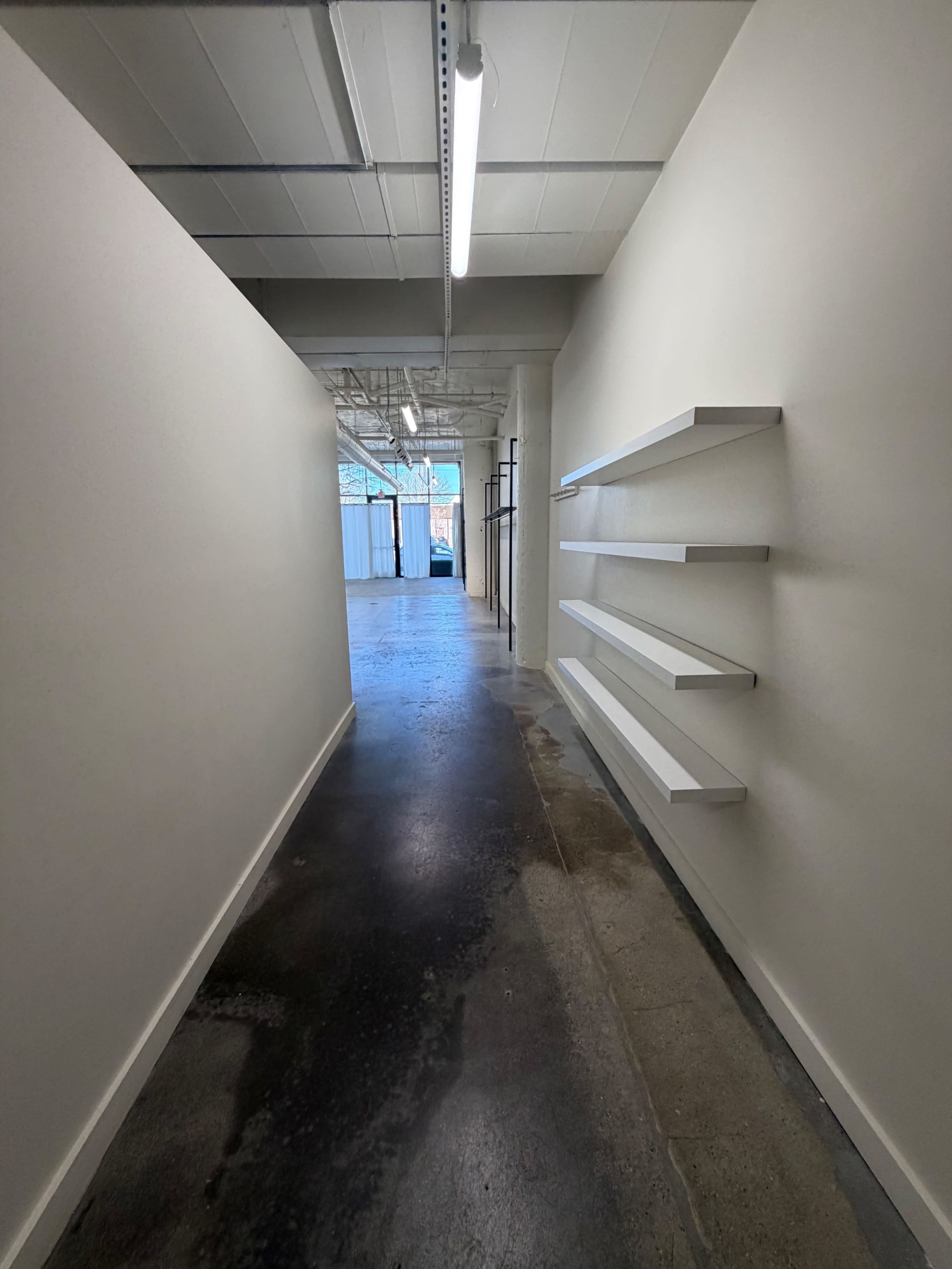 The image shows a narrow hallway with white walls and three mounted shelves on the right side, leading to an illuminated, open space at the end.