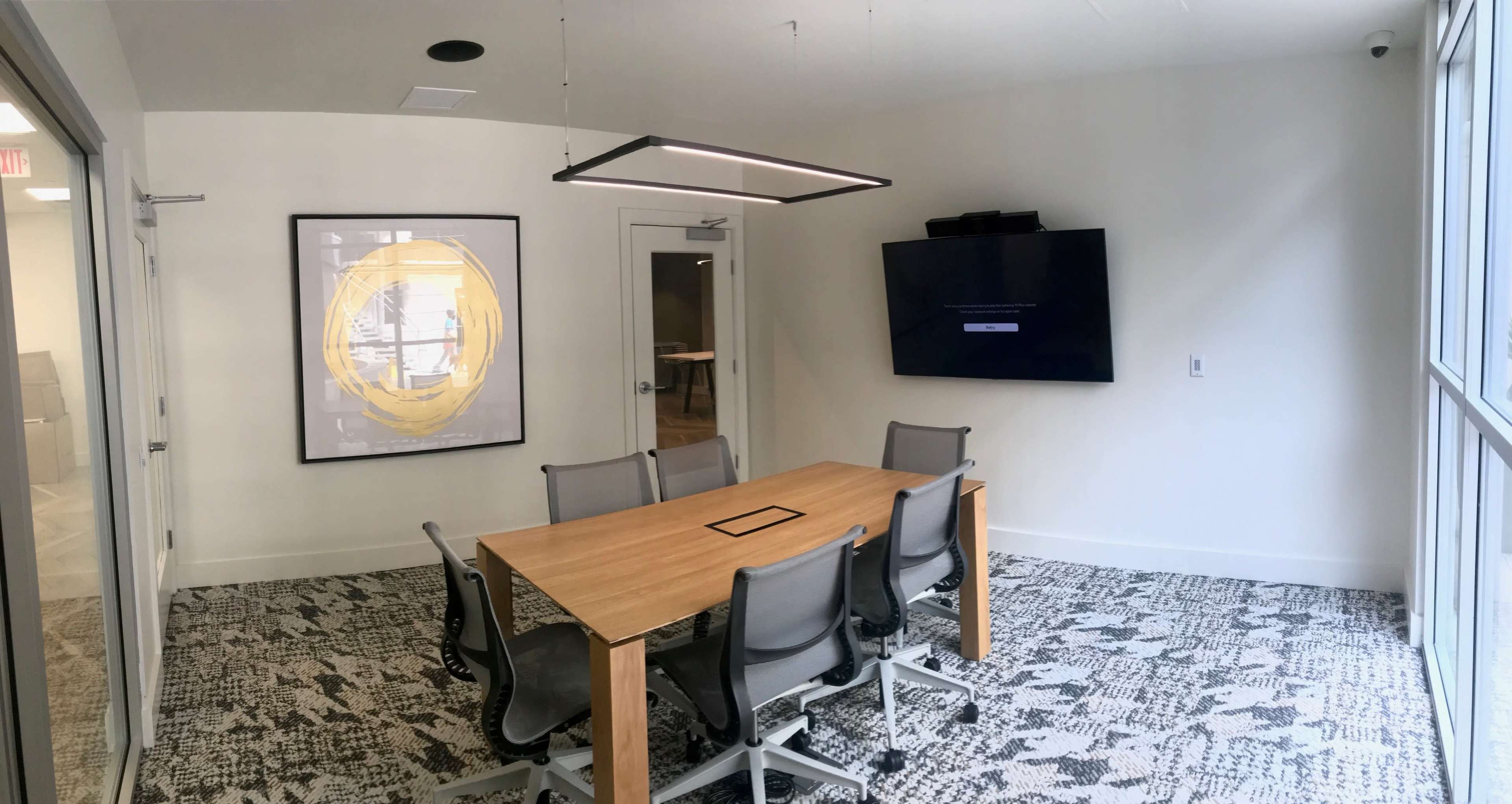 A conference room features a wooden table surrounded by six chairs, a wall-mounted screen, and a large framed artwork.