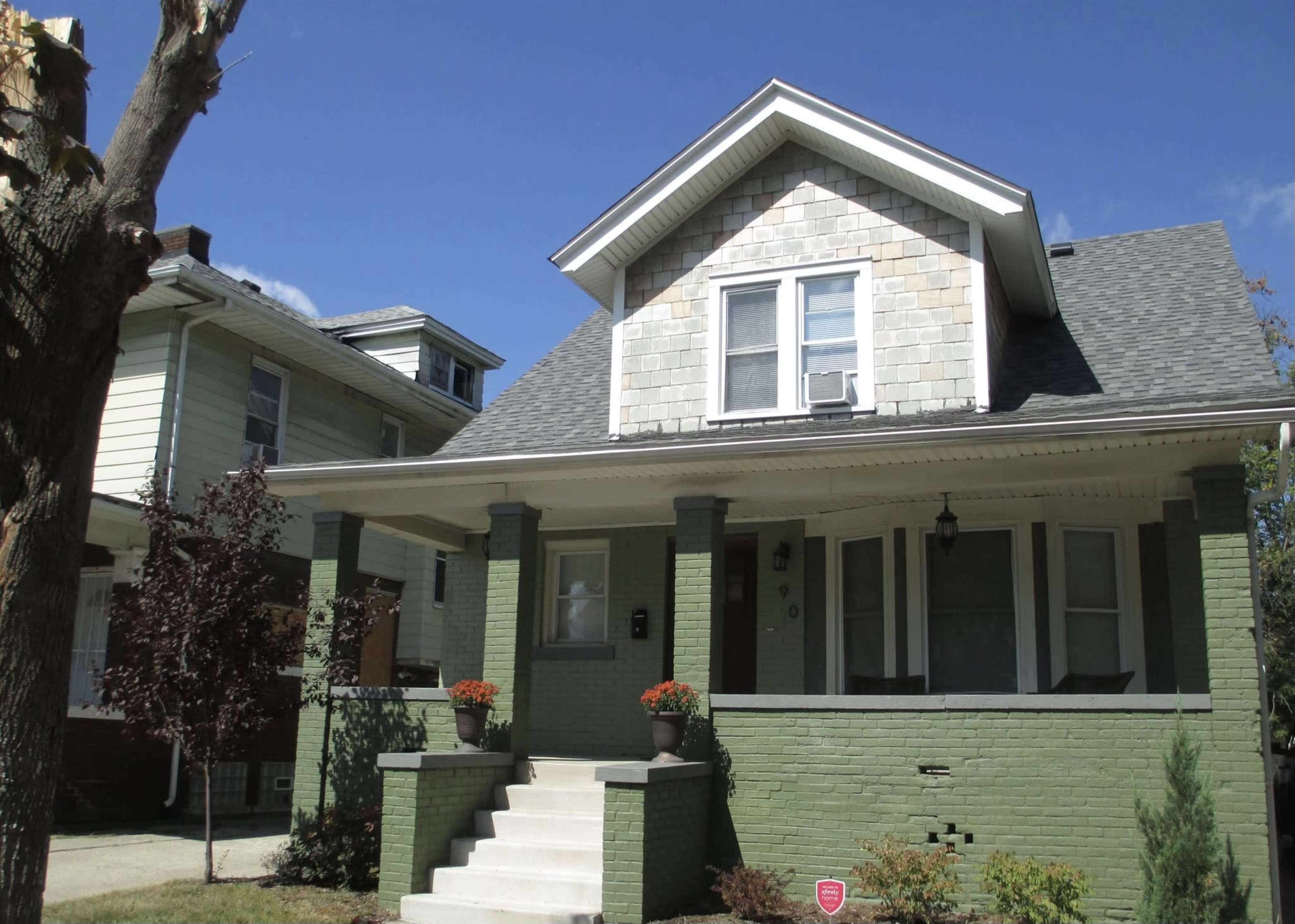 The image shows a two-story house with a green-painted lower level, a peaked roof, and stone-shingled upper siding, flanked by two smaller trees and a neighboring house.