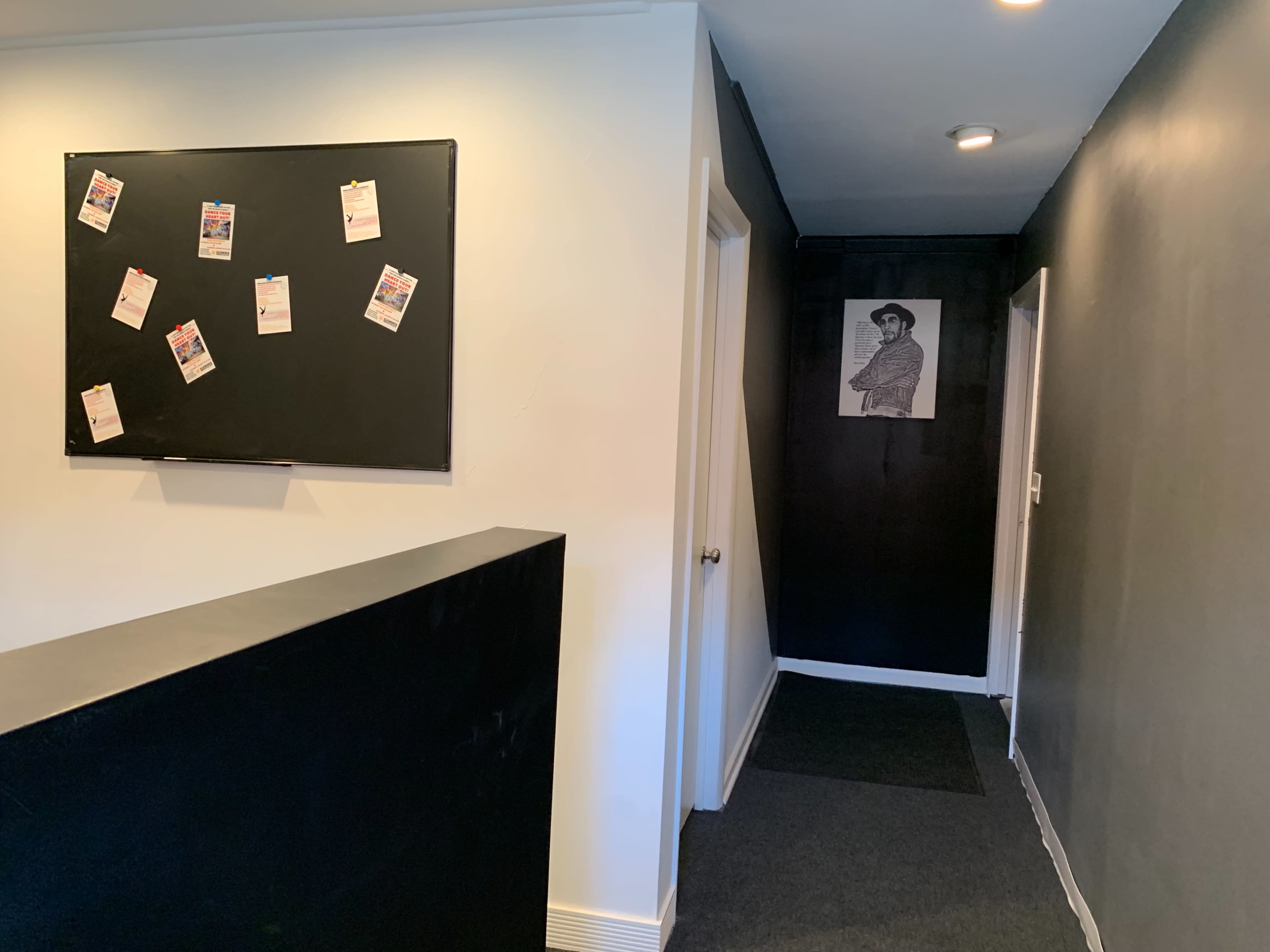A hallway with a black bulletin board on the left, a dark wall on the right, and a doorway at the end of the corridor.