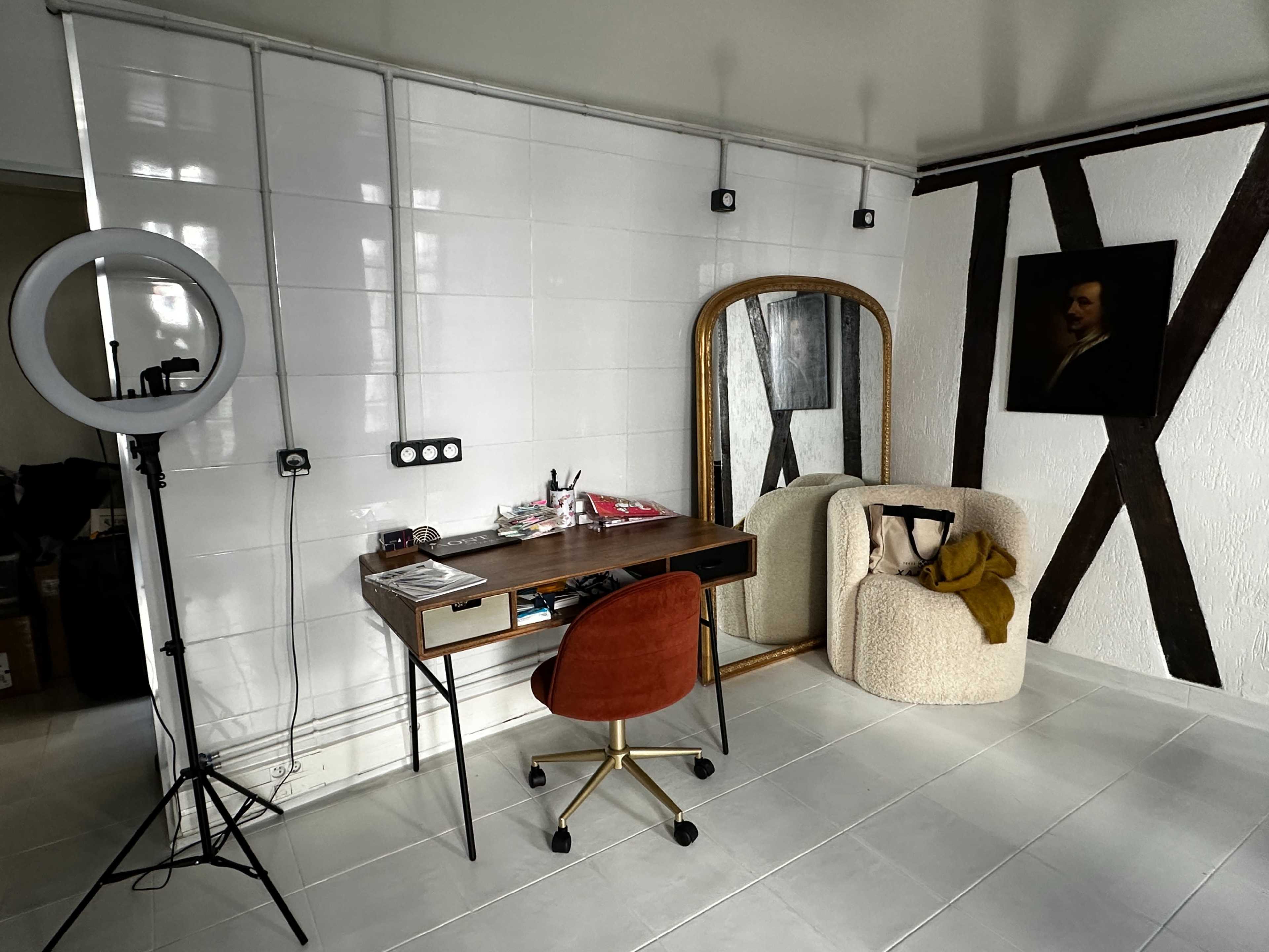 The image shows a modern workspace featuring a wooden desk with a chair, a large round mirror, and a cozy armchair, set against a backdrop of white tiles and exposed wooden beams.