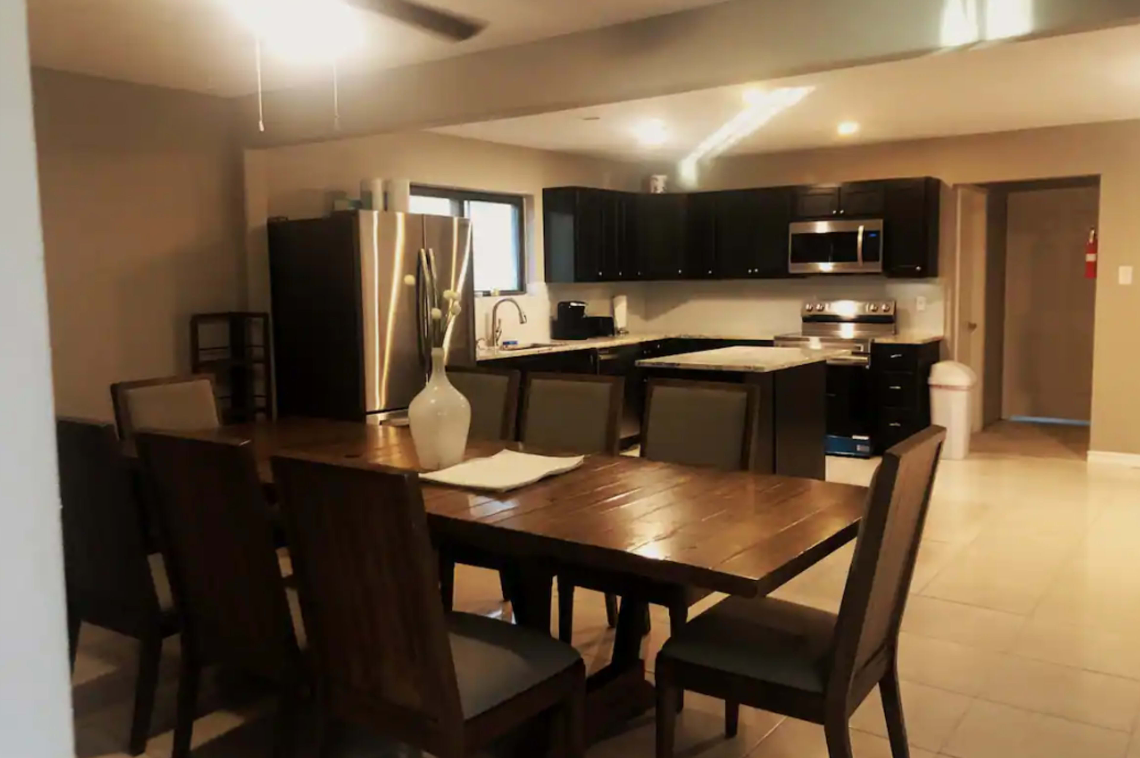 A modern kitchen and dining area featuring a wooden table with six chairs, stainless steel appliances, and dark cabinetry.
