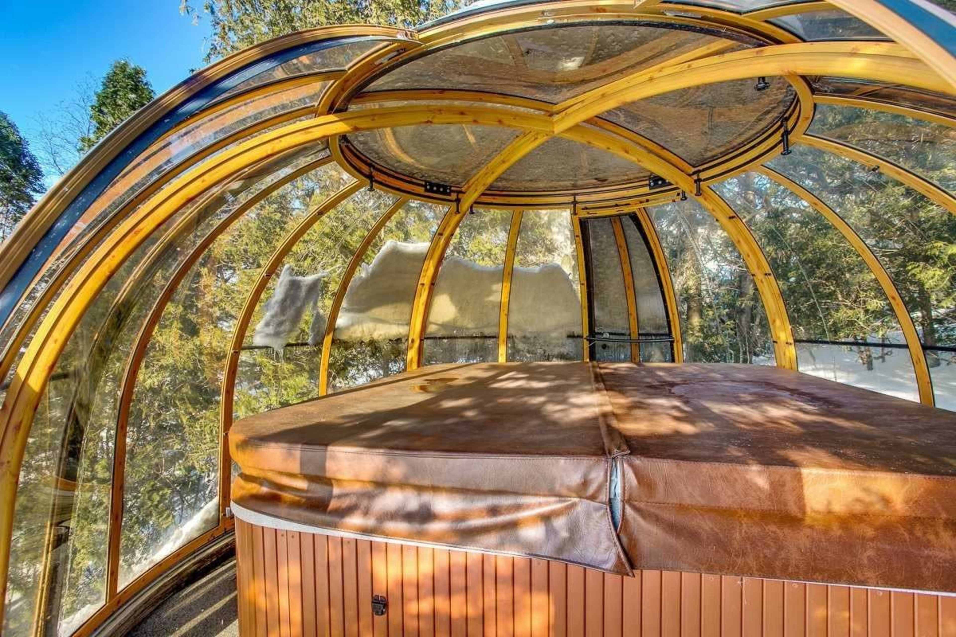 A circular wooden hot tub enclosed in a glass structure surrounded by trees.