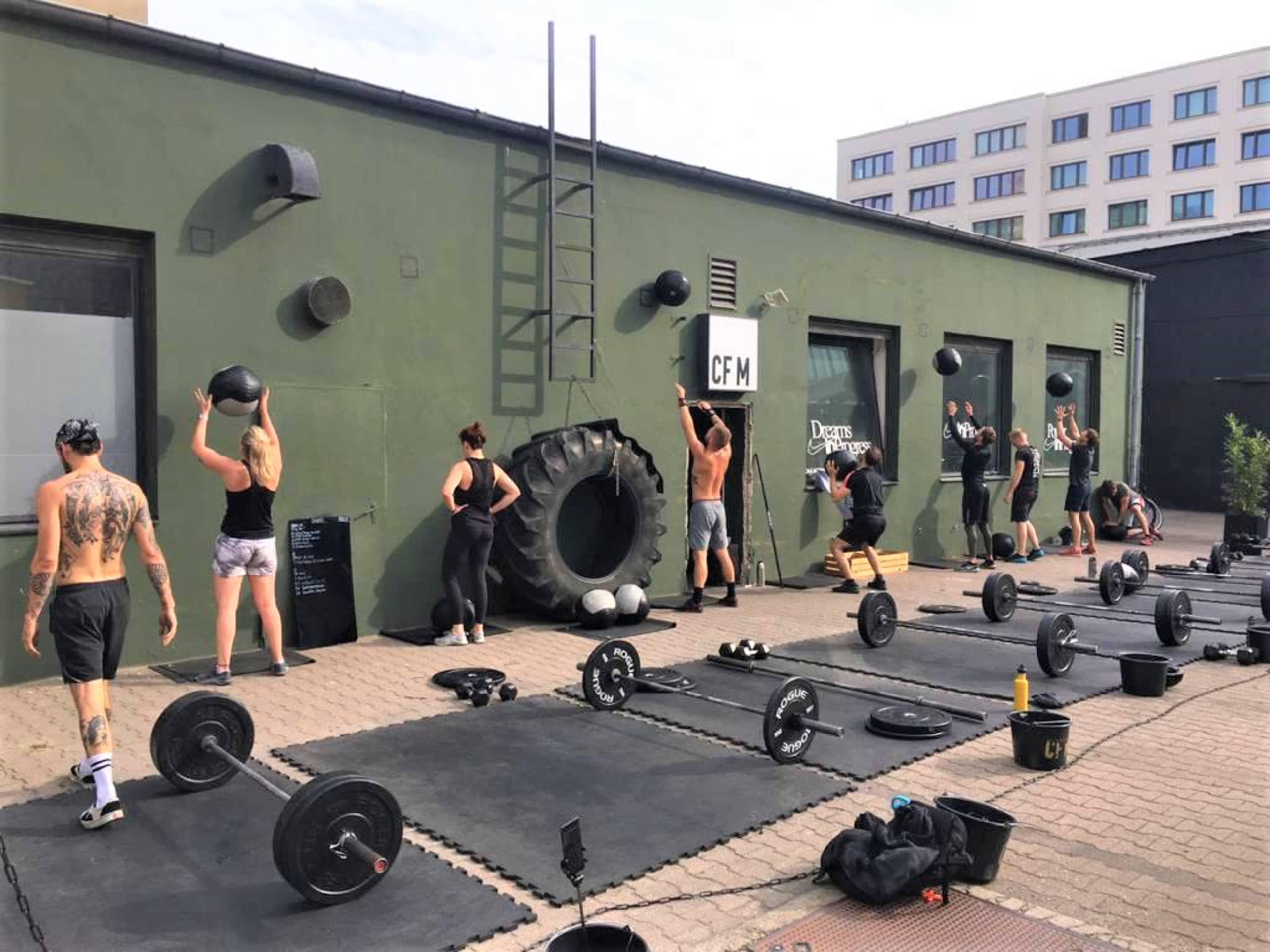 A group of people are participating in a workout session outside, using weights and medicine balls near a gym with a large tire and exercise mats.