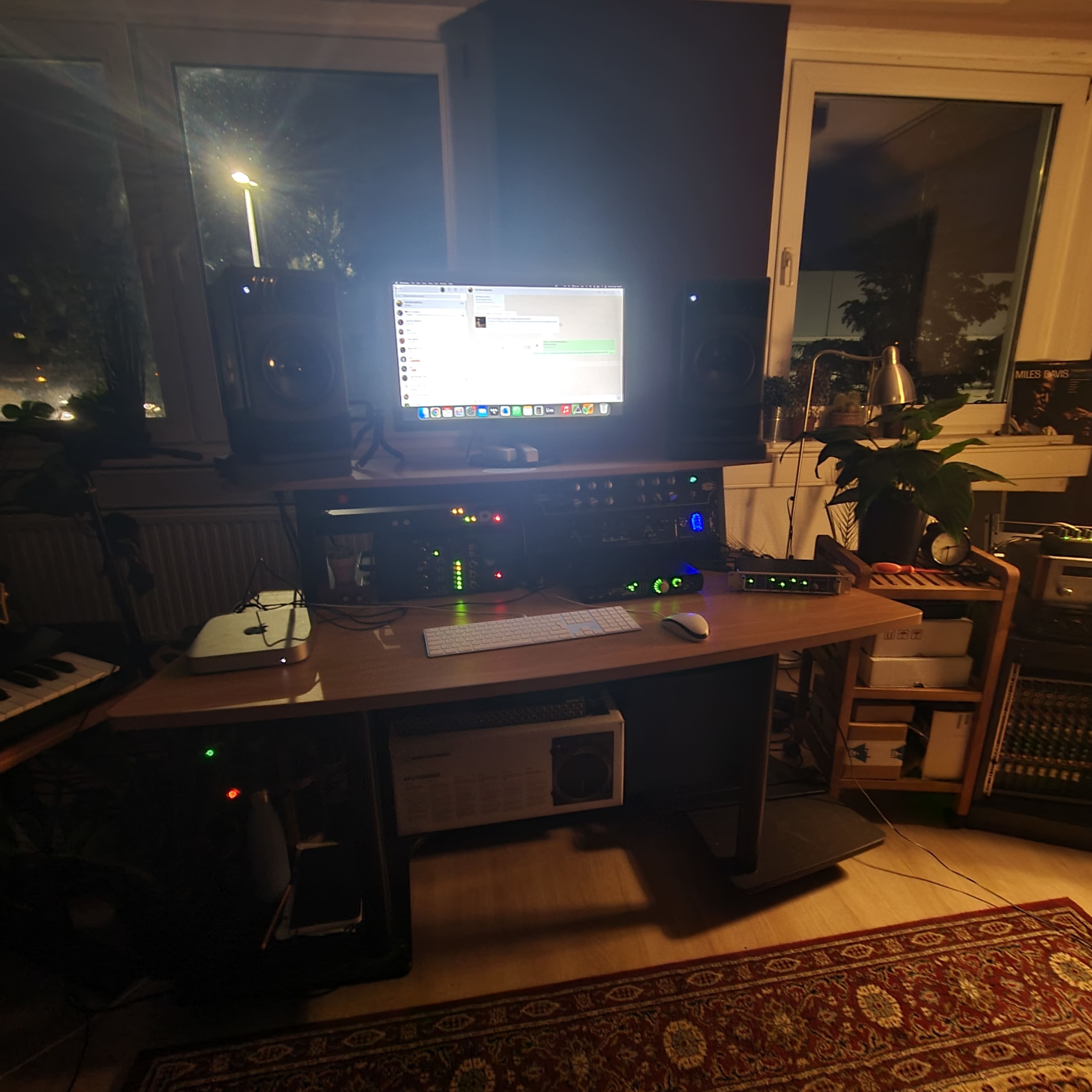 A computer workstation with studio monitors and audio equipment is set up in a room with large windows and plants.