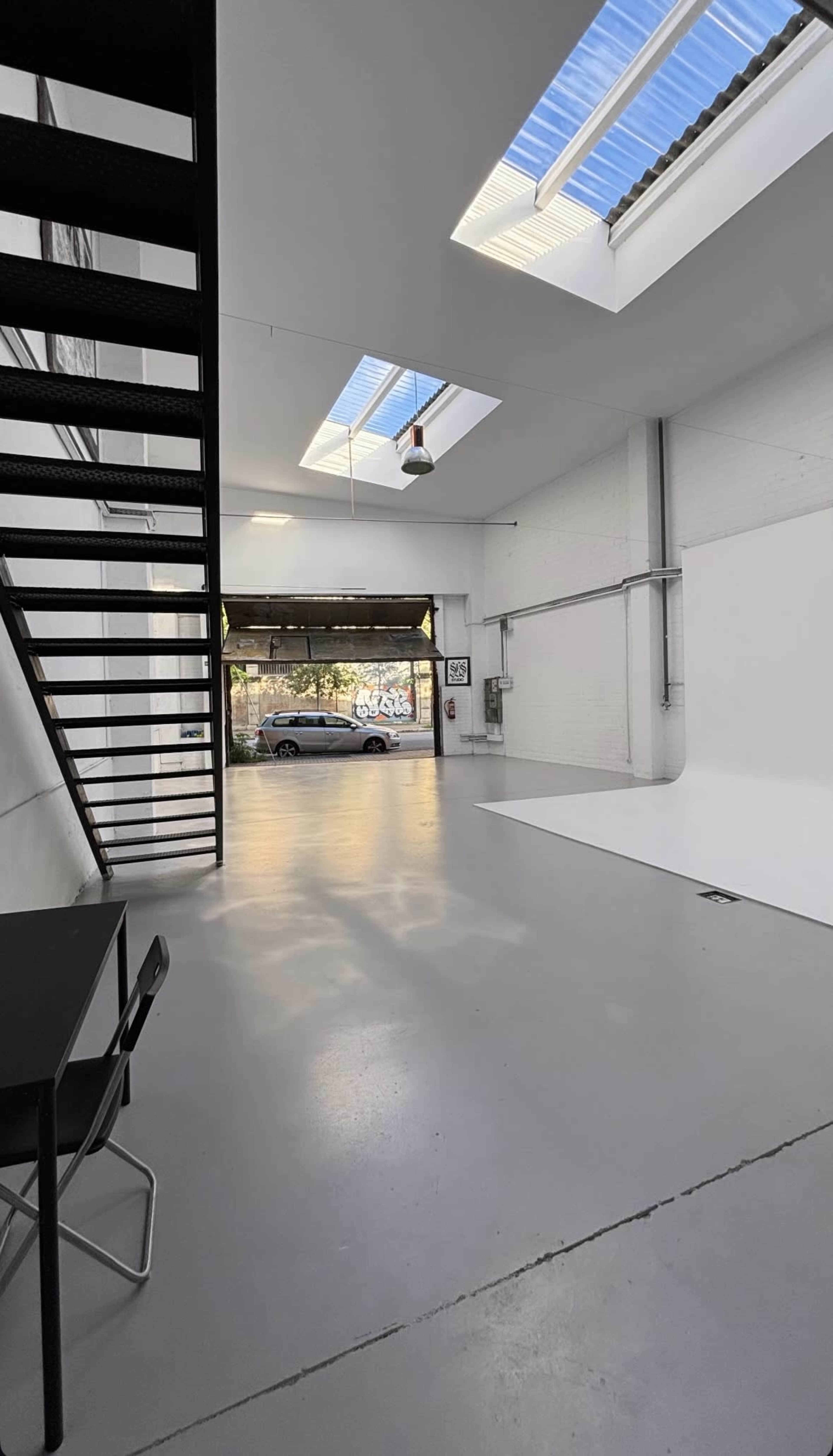 The image shows an empty studio space with a white backdrop, large skylights, and a staircase leading to an upper level.