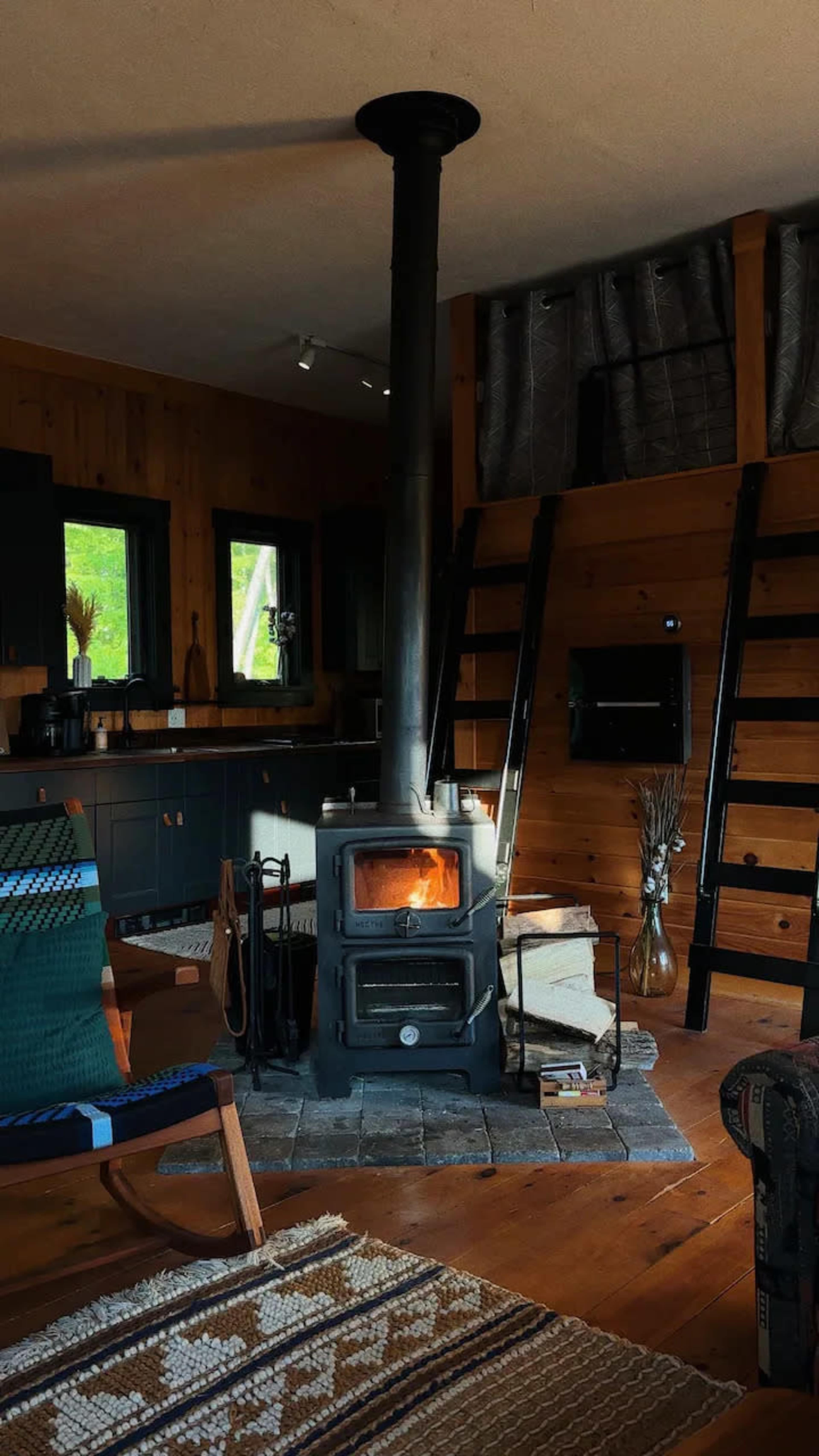 A wood stove stands centrally in a rustic cabin living area, surrounded by wooden walls and a patterned rug.