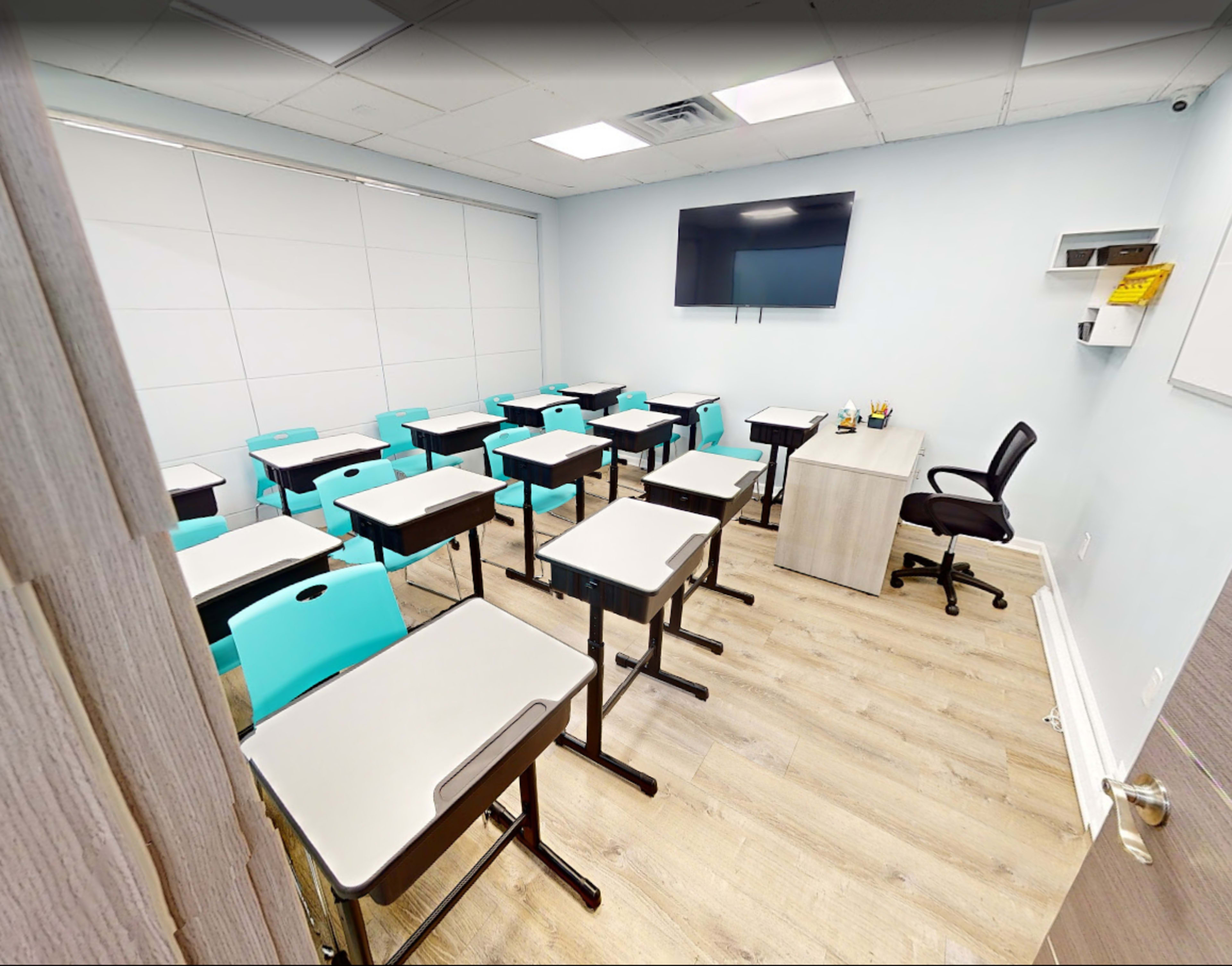 The image shows a well-organized classroom with plastic desks and chairs arranged in neat rows, a wall-mounted television, and a wooden teacher's desk.