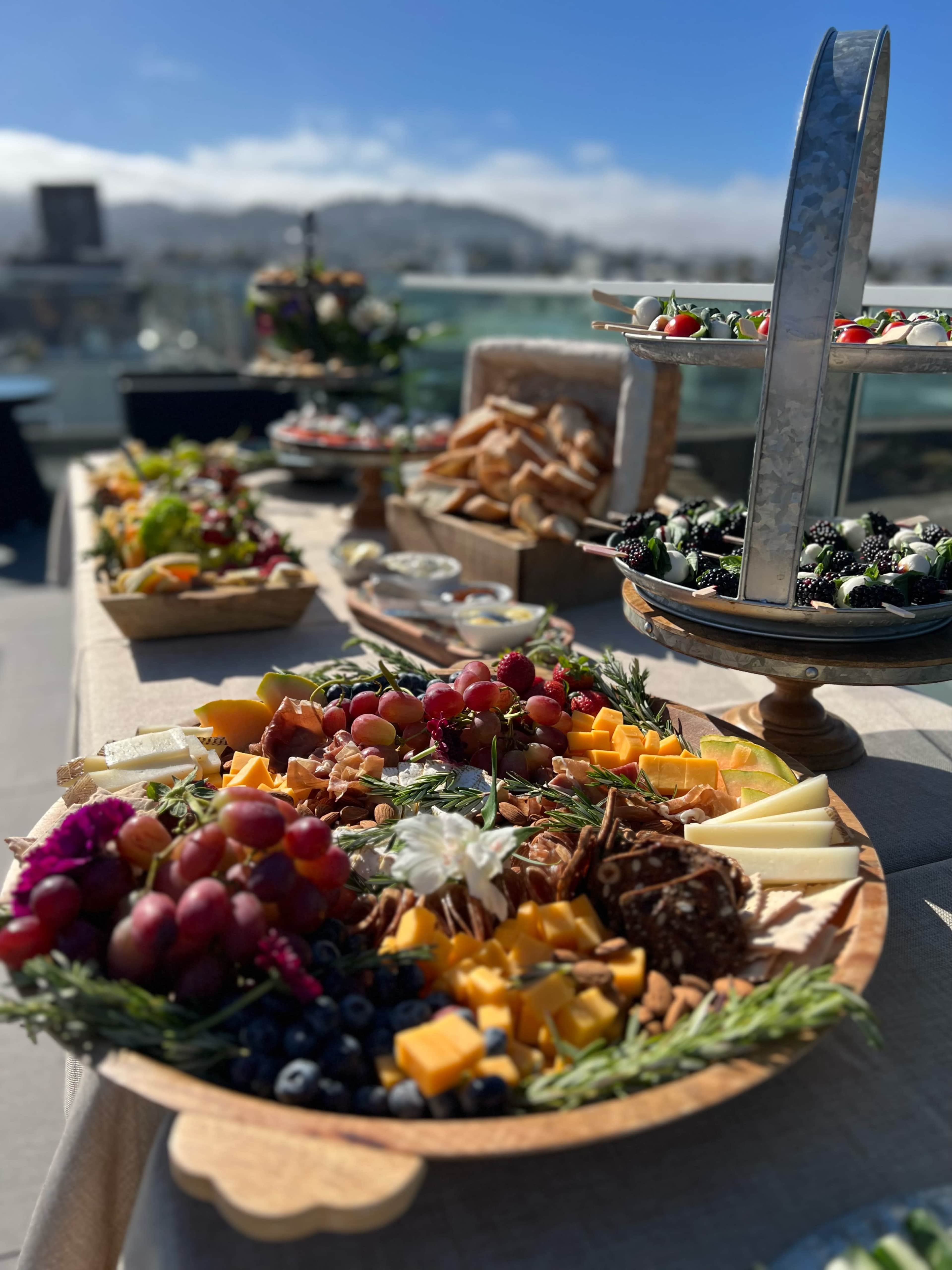 A large wooden platter filled with assorted cheeses, fruits, nuts, and garnishes, set against a backdrop of a sunny outdoor venue.