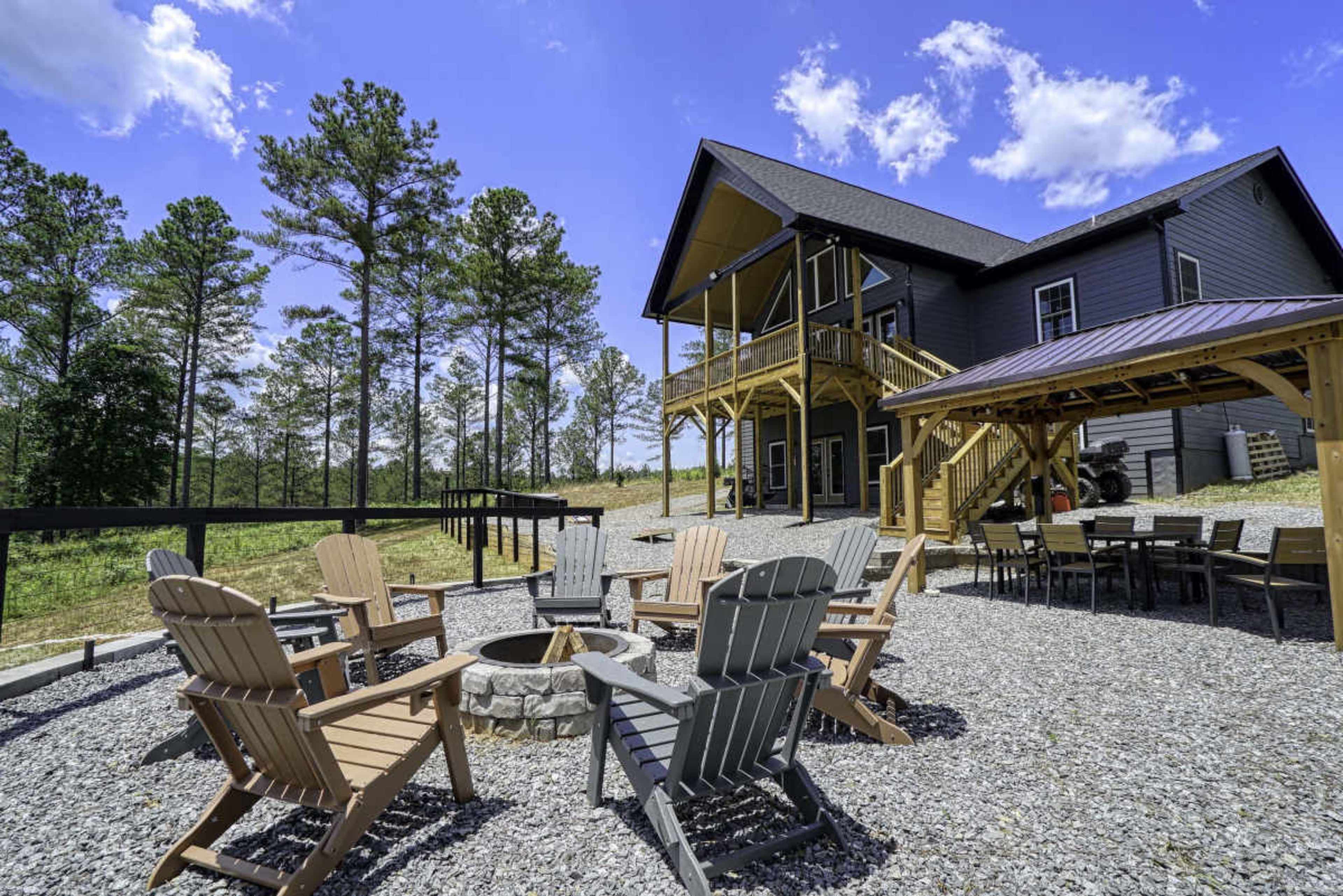 A spacious outdoor area features a fire pit surrounded by wooden Adirondack chairs, with a large house and covered seating area in the background.