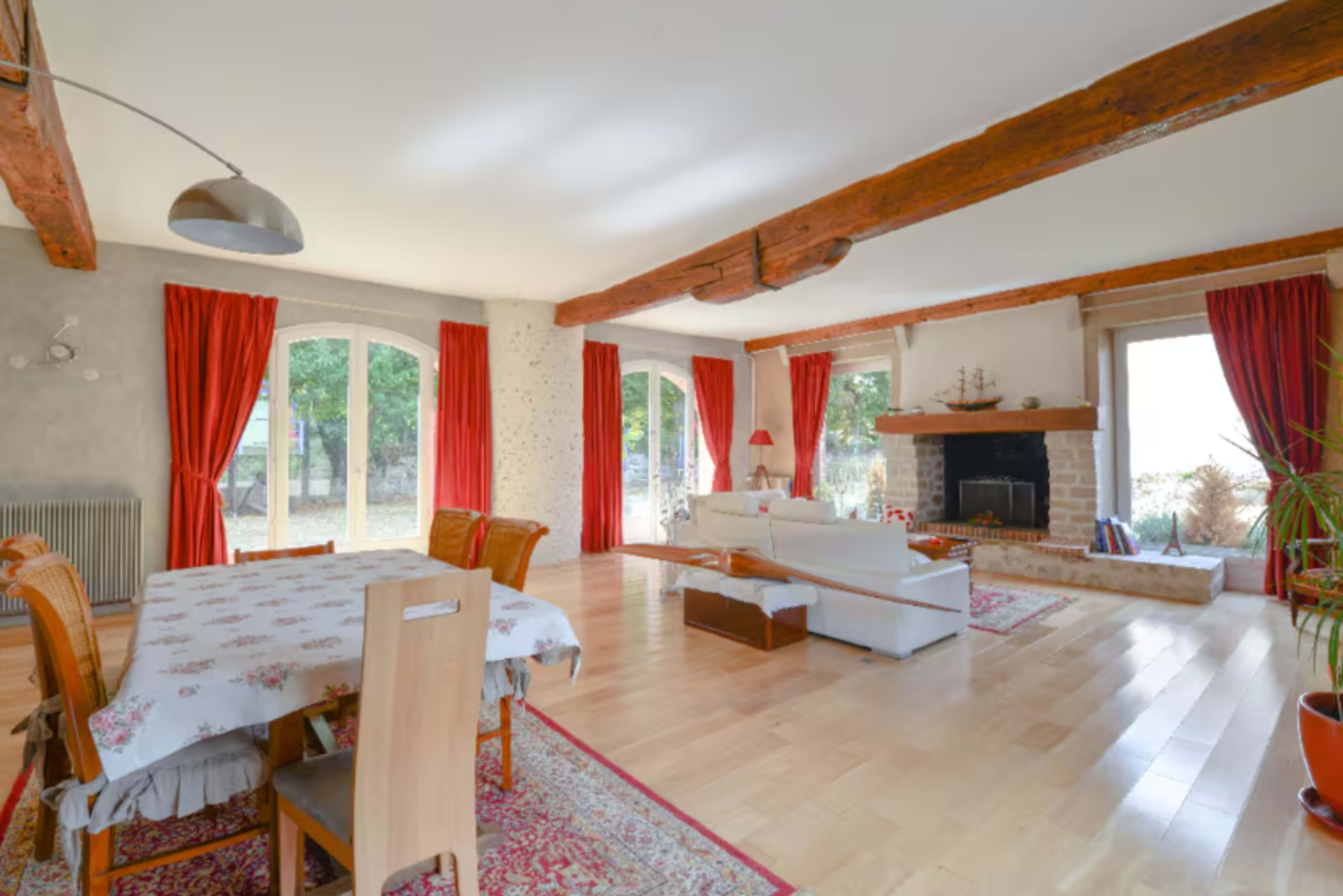 A spacious living area features a dining table, a white sofa, a fireplace, and large windows with red curtains.