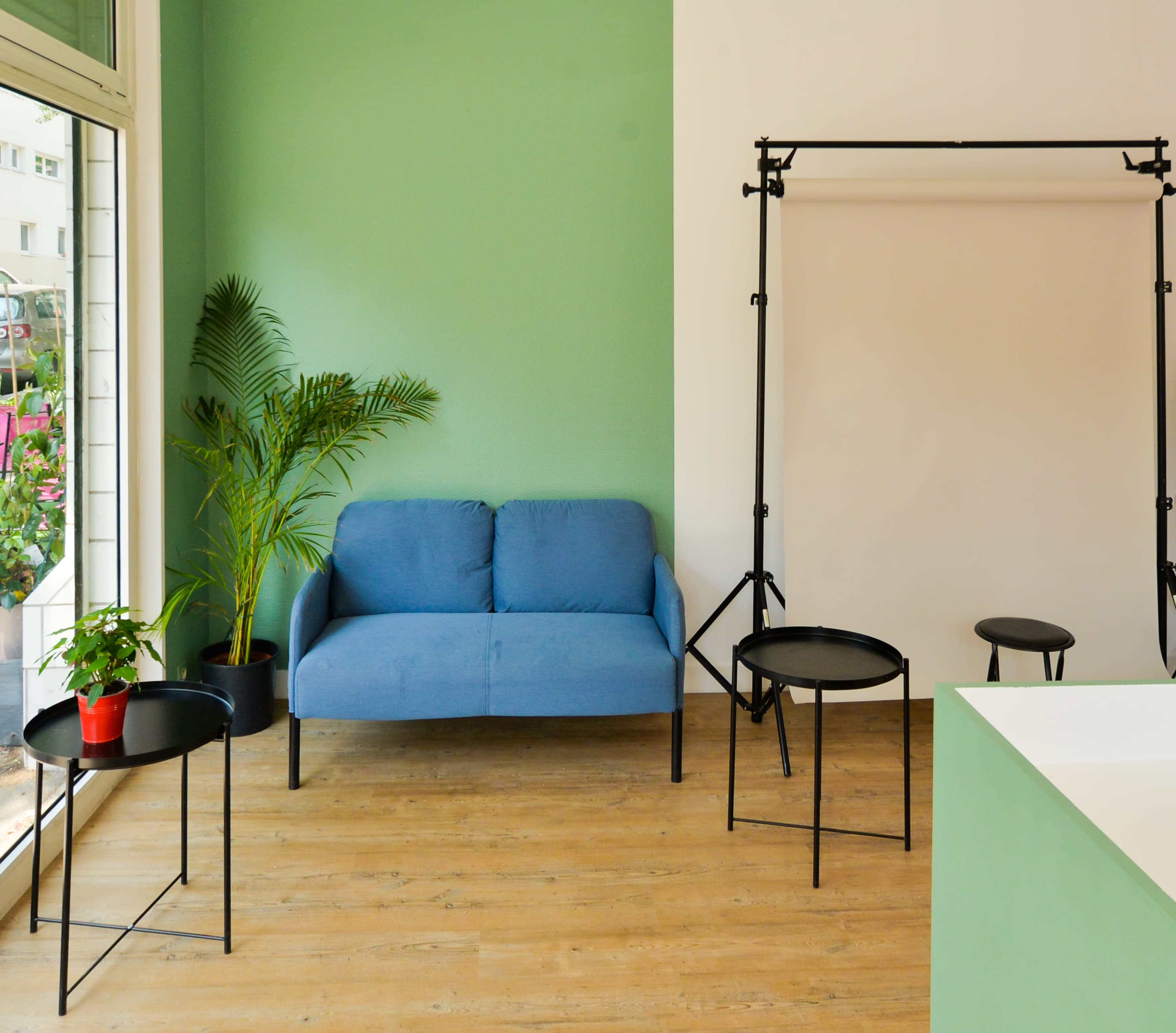 A minimalistic interior space featuring a blue couch, black side tables, a stool, and a potted plant against a green wall.
