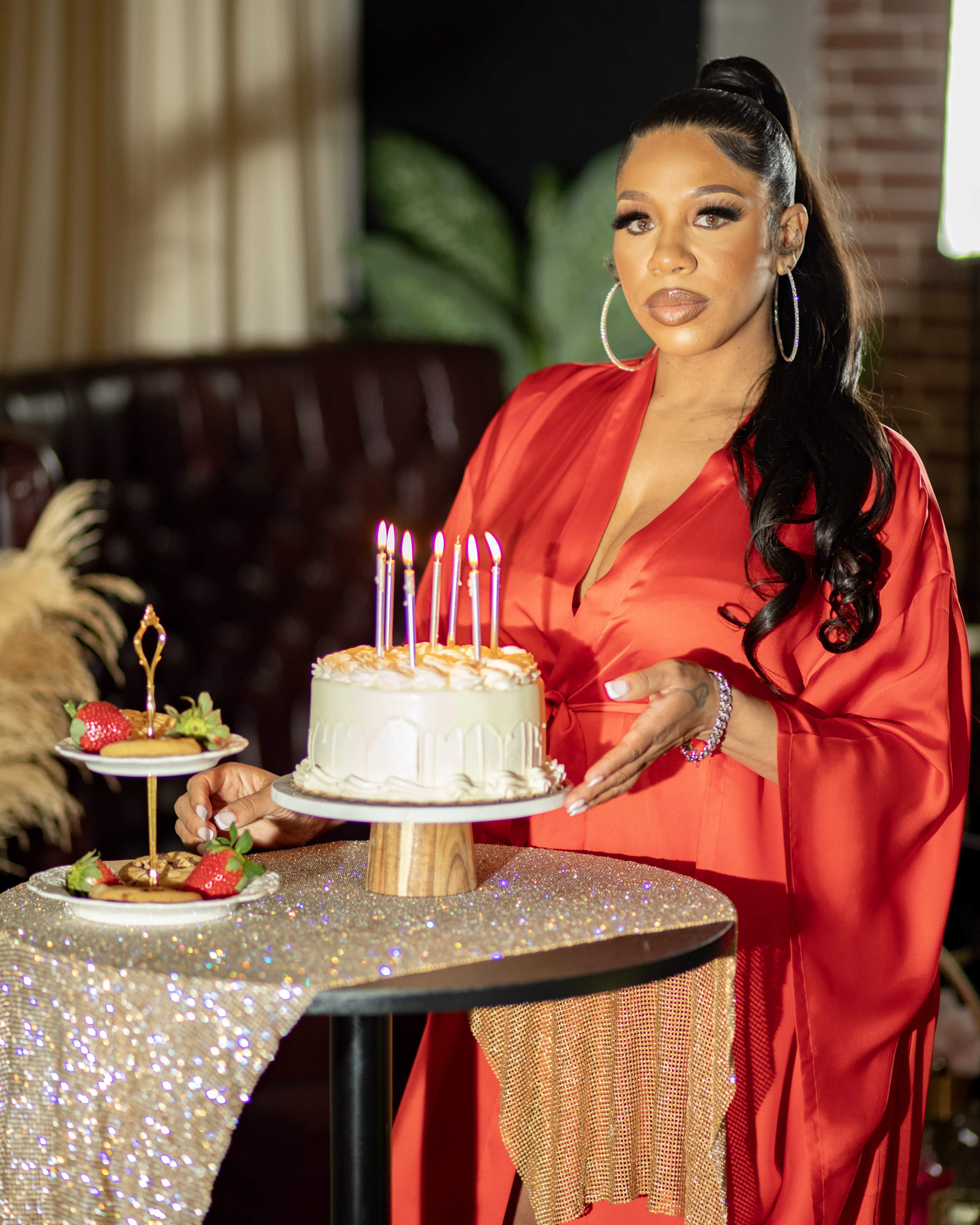 A woman in a red dress holds a decorated cake with candles on a table beside a platter of desserts.