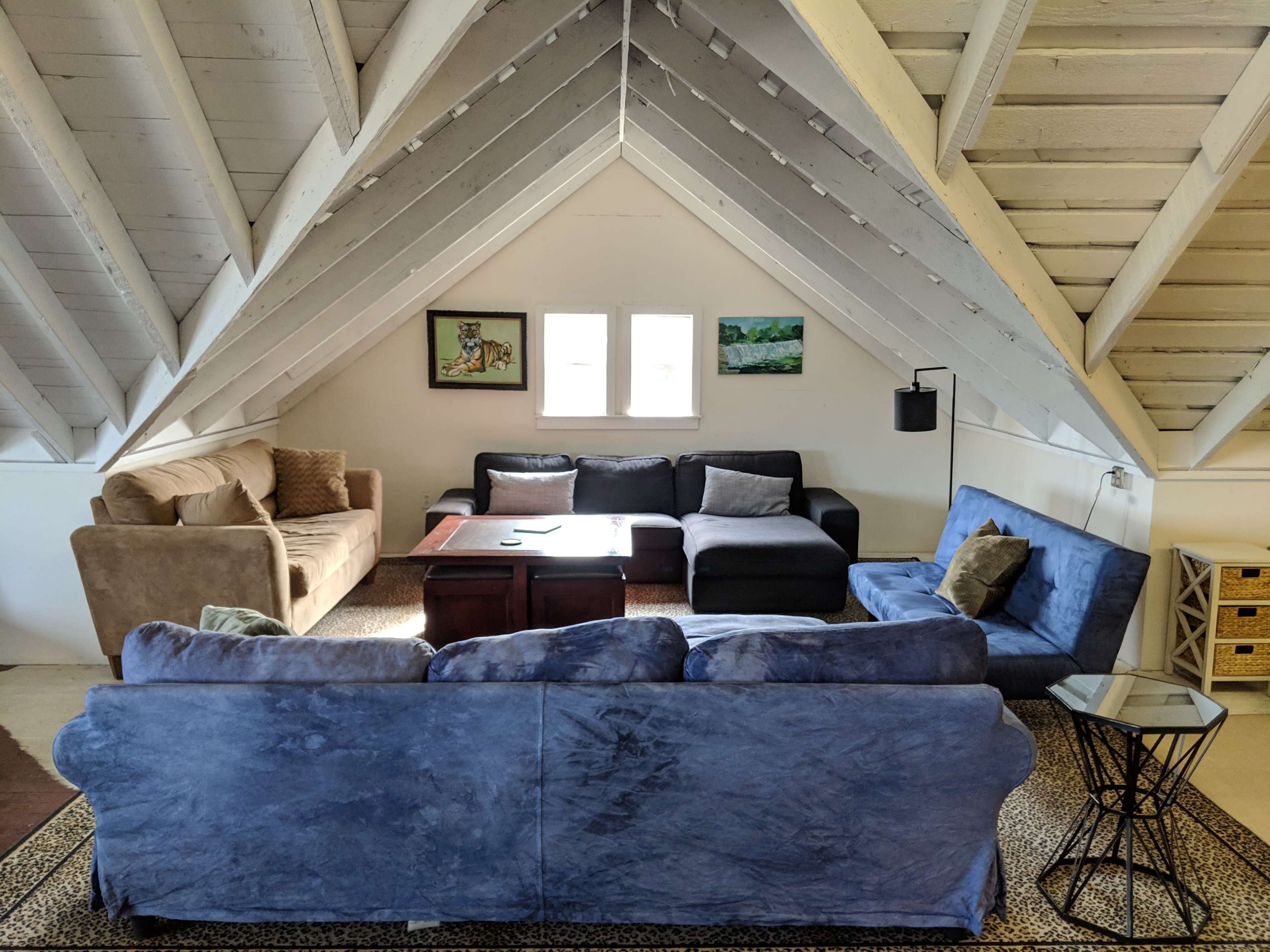 The scene features a spacious, sloped-ceiling living area with several sofas, a coffee table, and large windows allowing natural light.