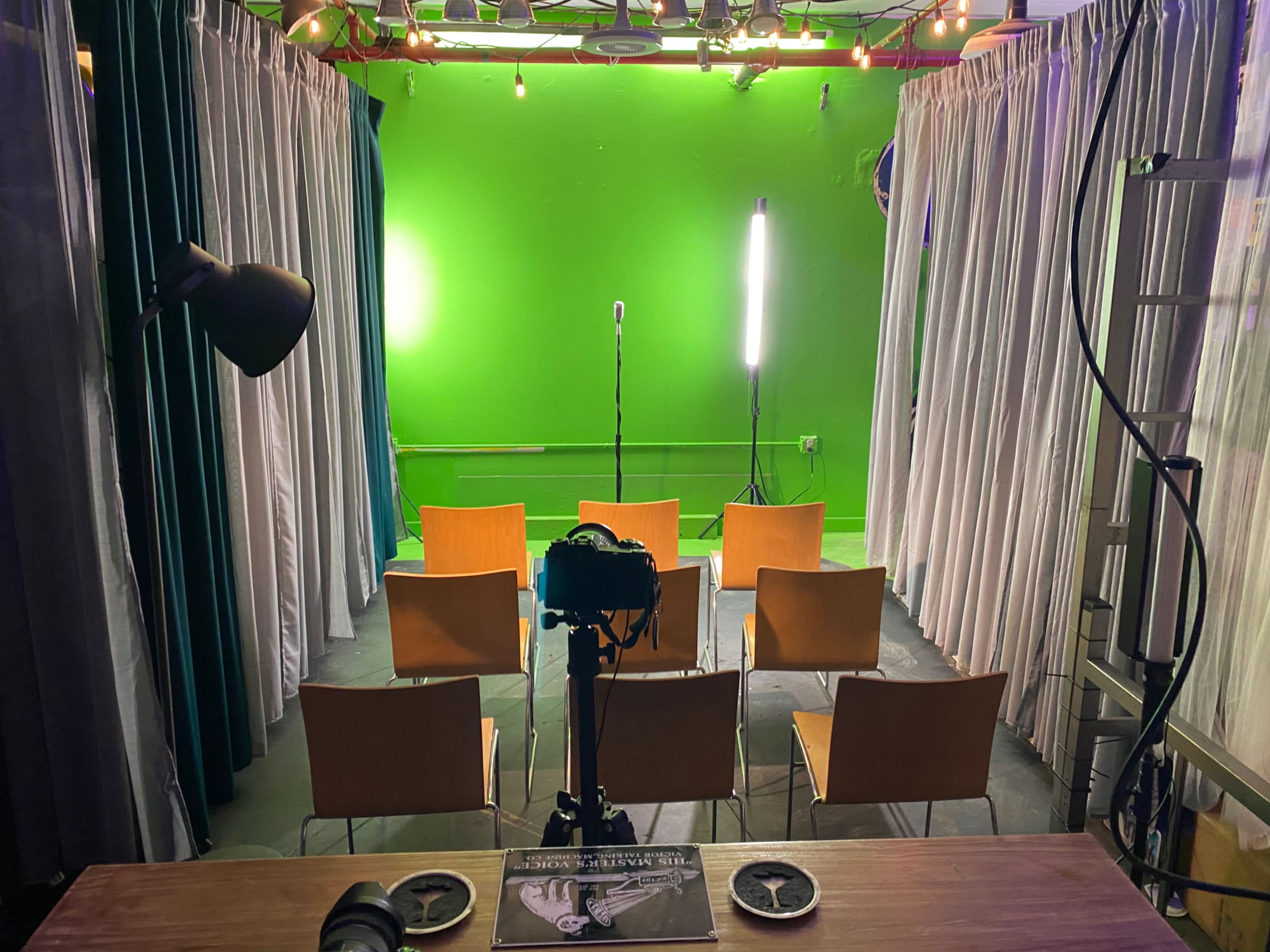 A small stage area with a microphone in front of a bright green backdrop, surrounded by empty wooden chairs and a camera on a table.