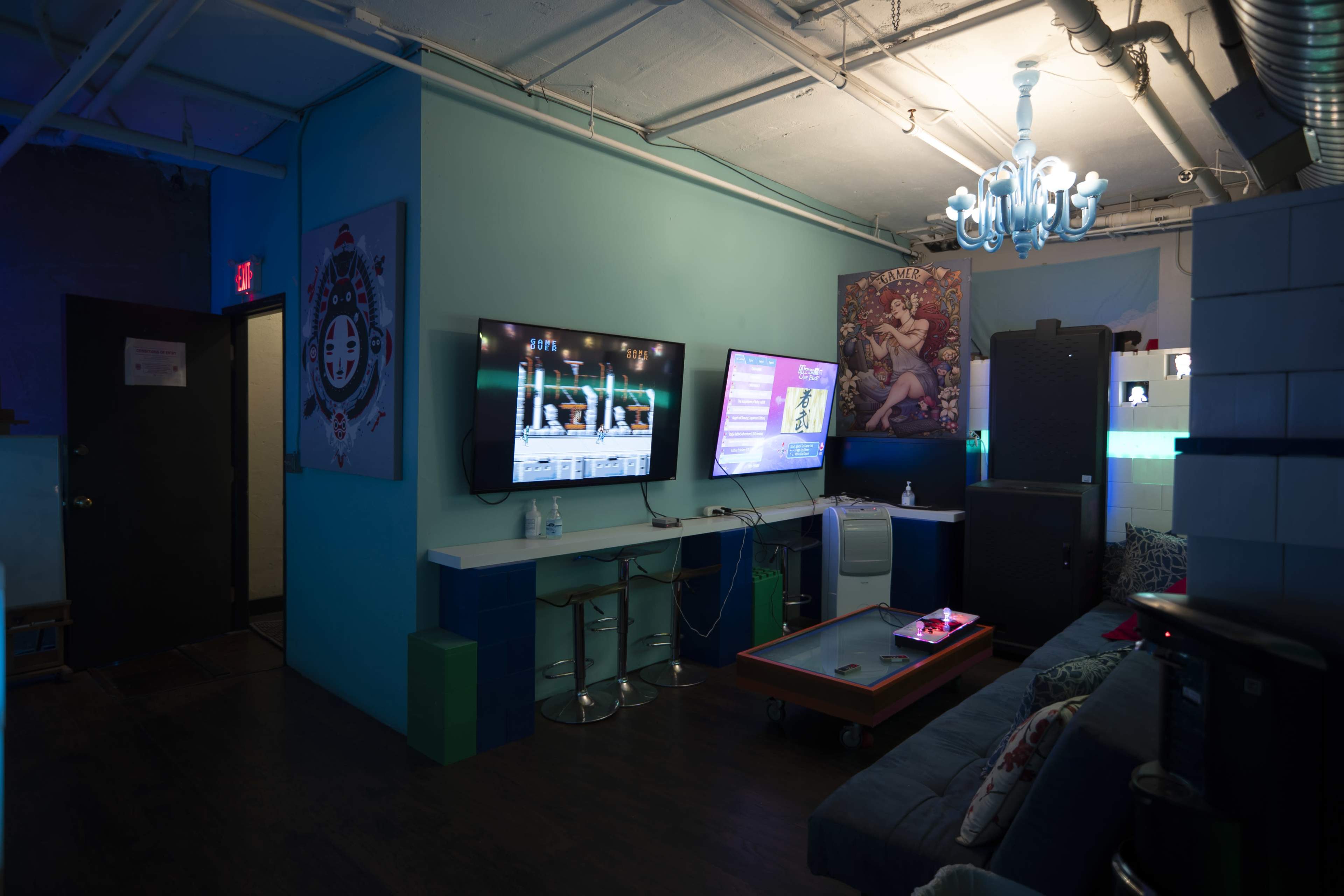 A dimly lit lounge area with two televisions on the wall, a coffee table in the center, and a mix of modern furniture and artwork.