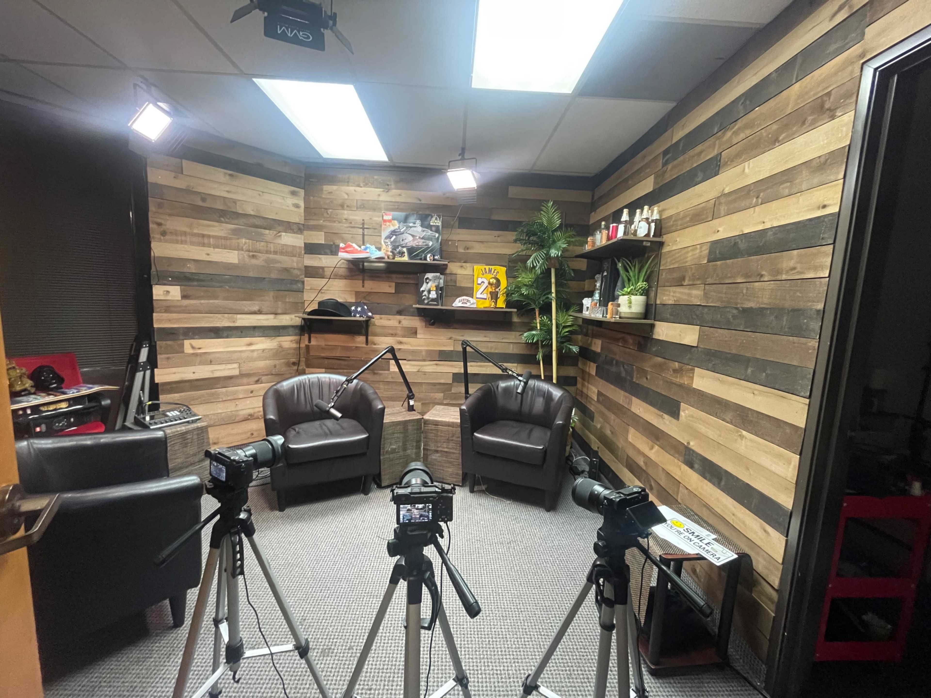 A recording setup in a room with wooden walls, featuring two black chairs, three cameras on tripods, and various decorative items on shelves.