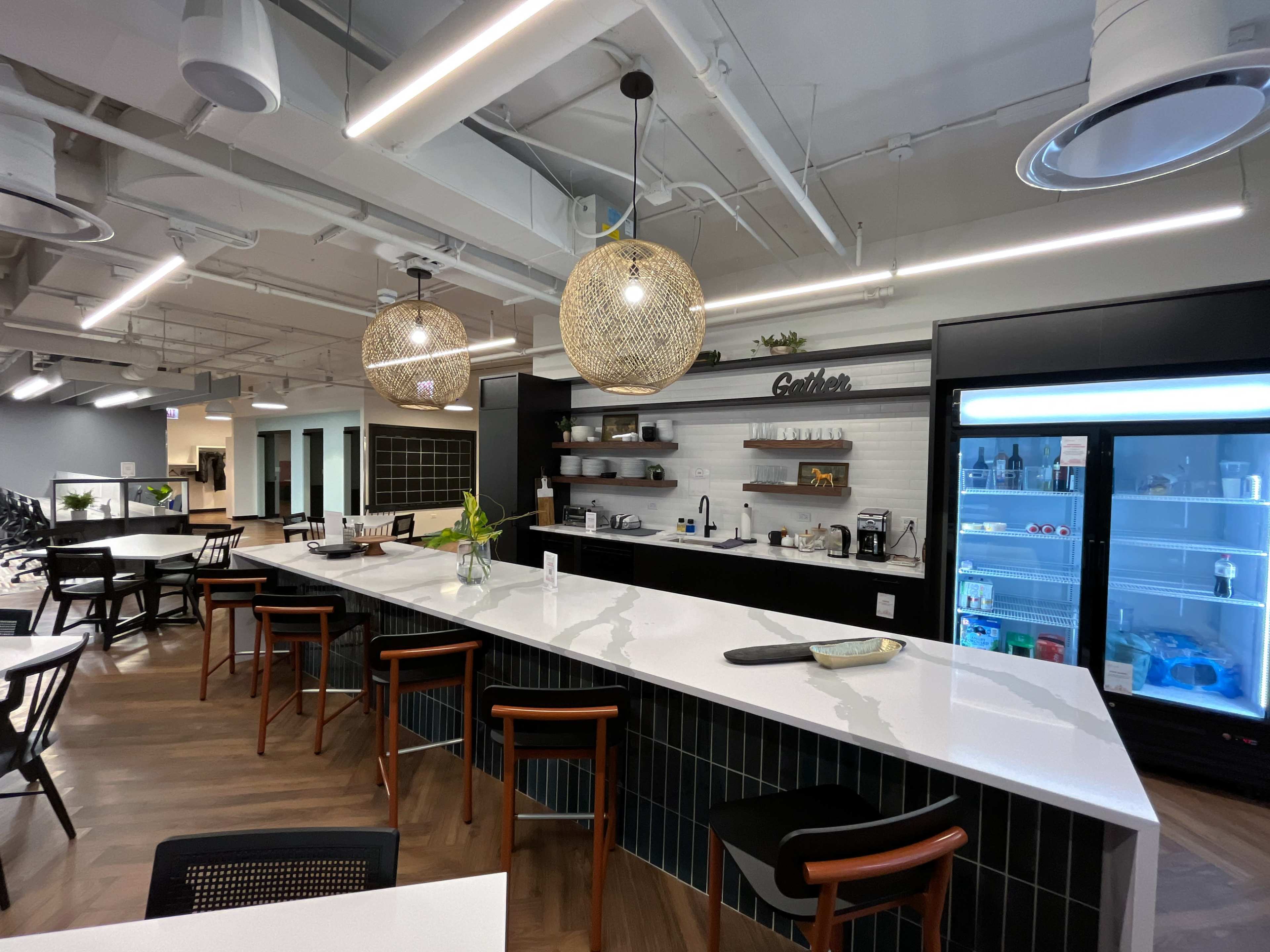 The image shows a modern kitchen area with a large white countertop, wooden bar stools, decorative light fixtures, and a fridge featuring a variety of beverages.