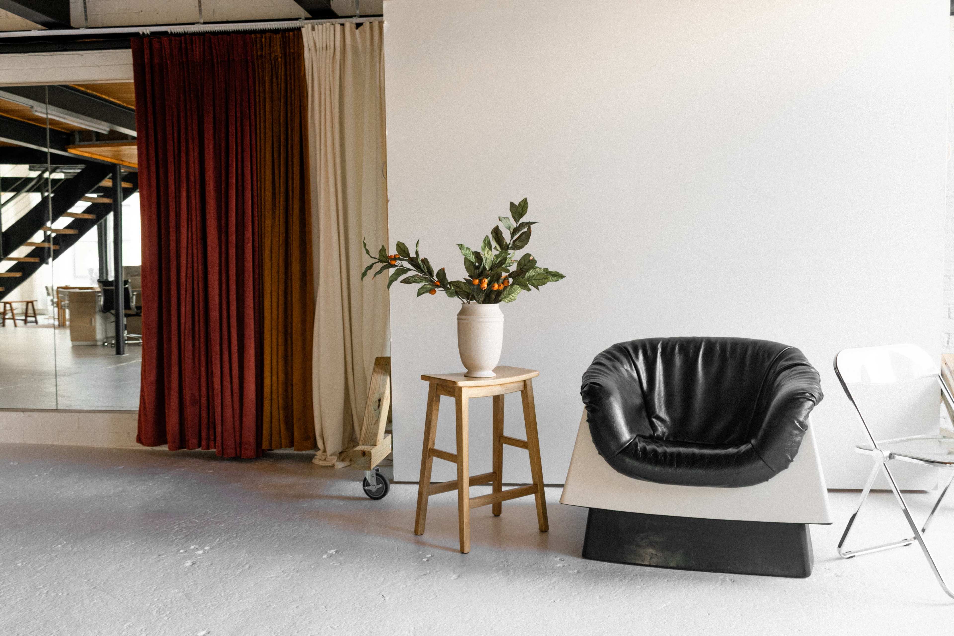 The scene features a black chair beside a wooden stool with a potted plant, framed by curtains of varying colors, and an open space leading to a staircase.
