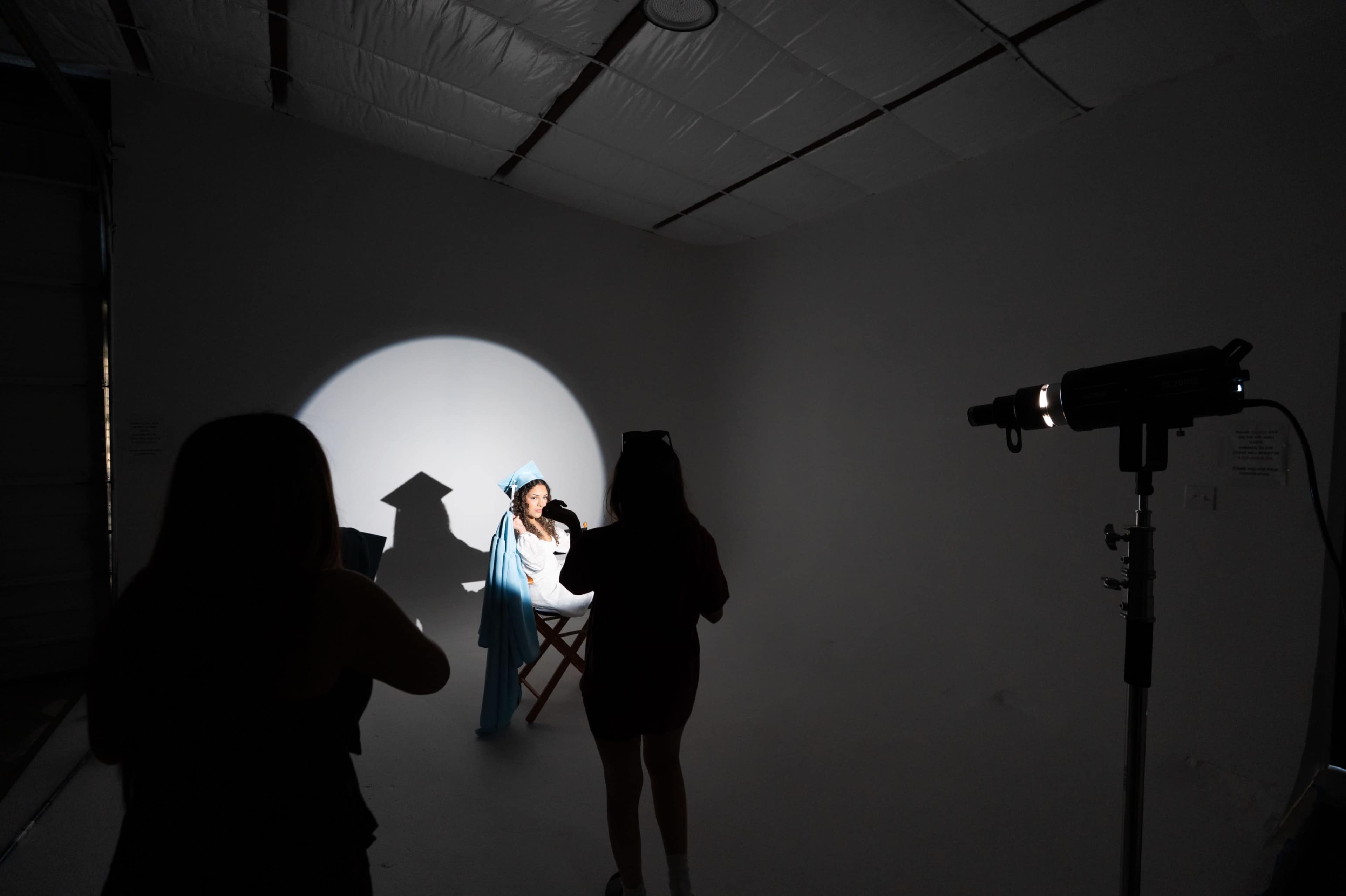 A woman seated in a chair is illuminated by a spotlight in a minimalistic studio while another person stands nearby, holding a camera.