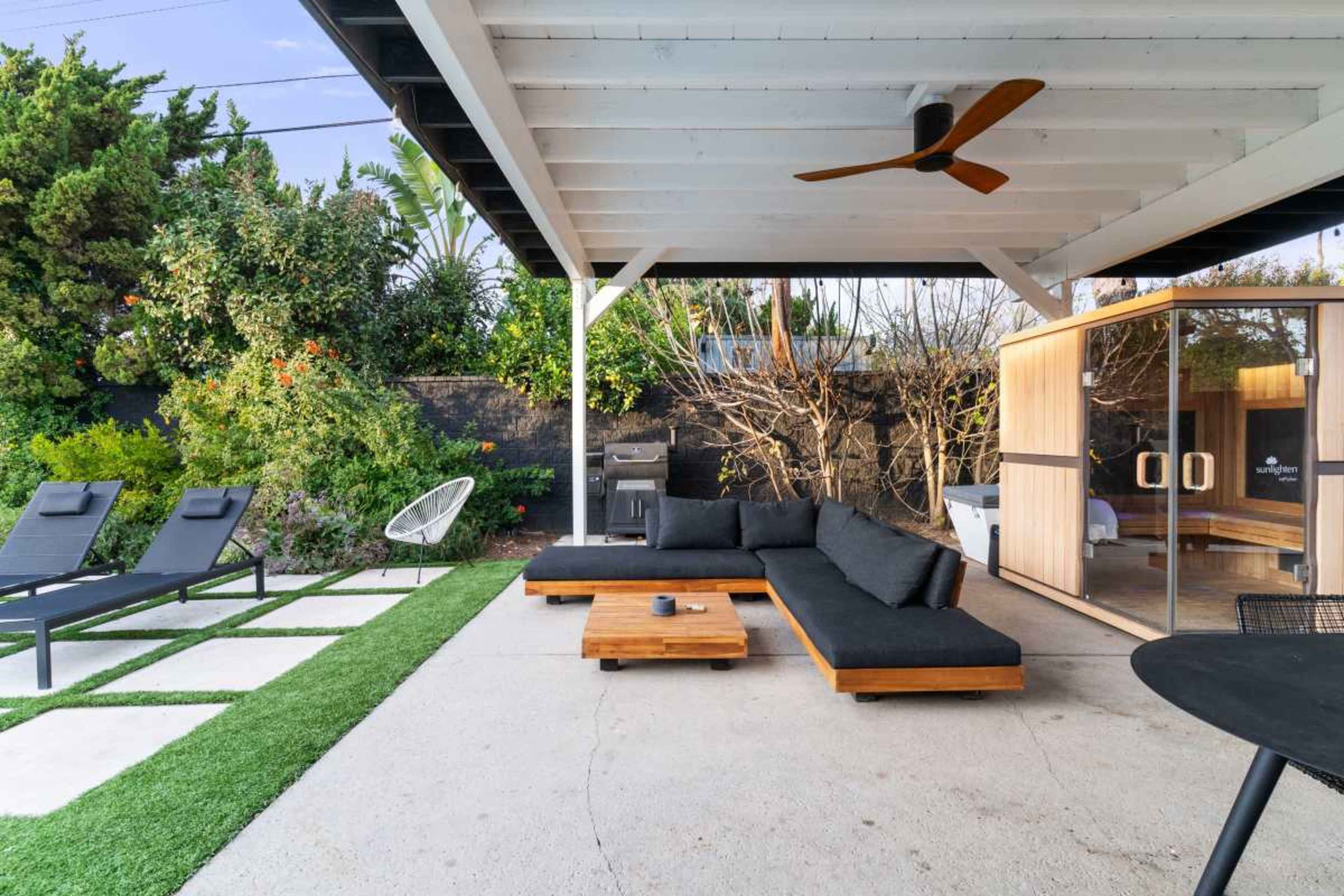 The image shows a modern outdoor patio featuring a sectional sofa, a coffee table, lounge chairs, and a grill surrounded by greenery.