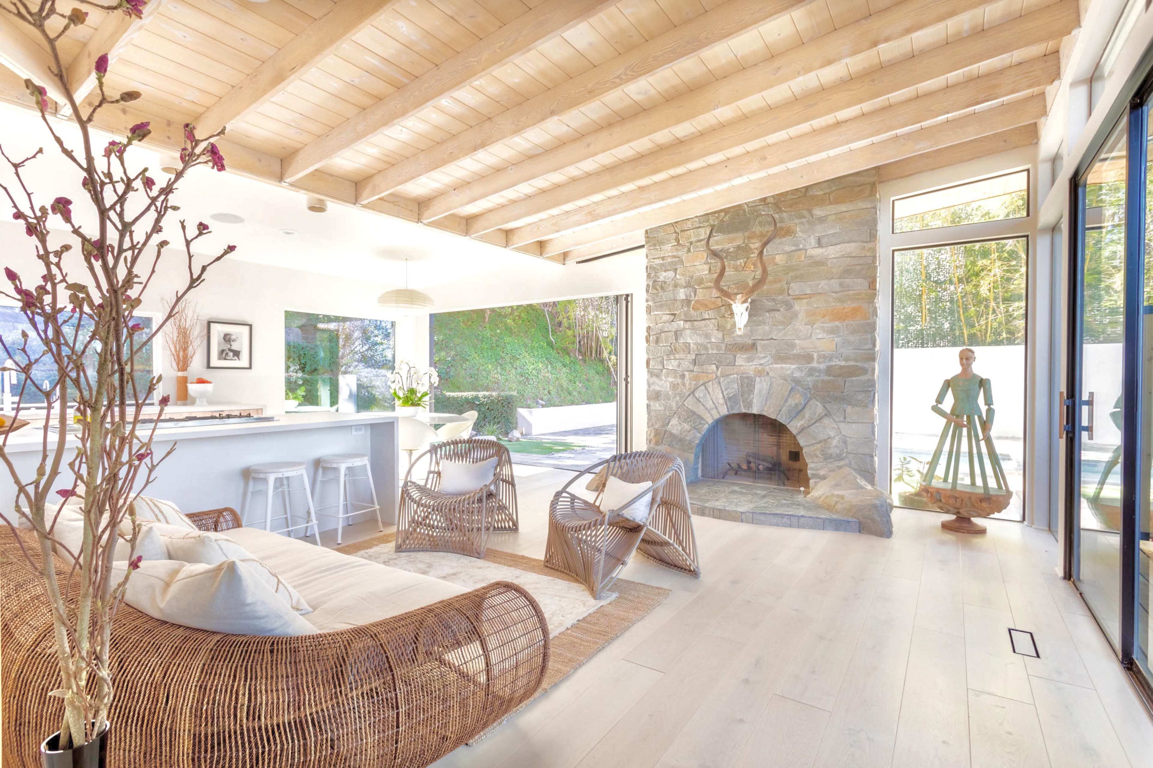 A modern living room with a stone fireplace, wooden ceiling beams, and large windows overlooking a green outdoor space.