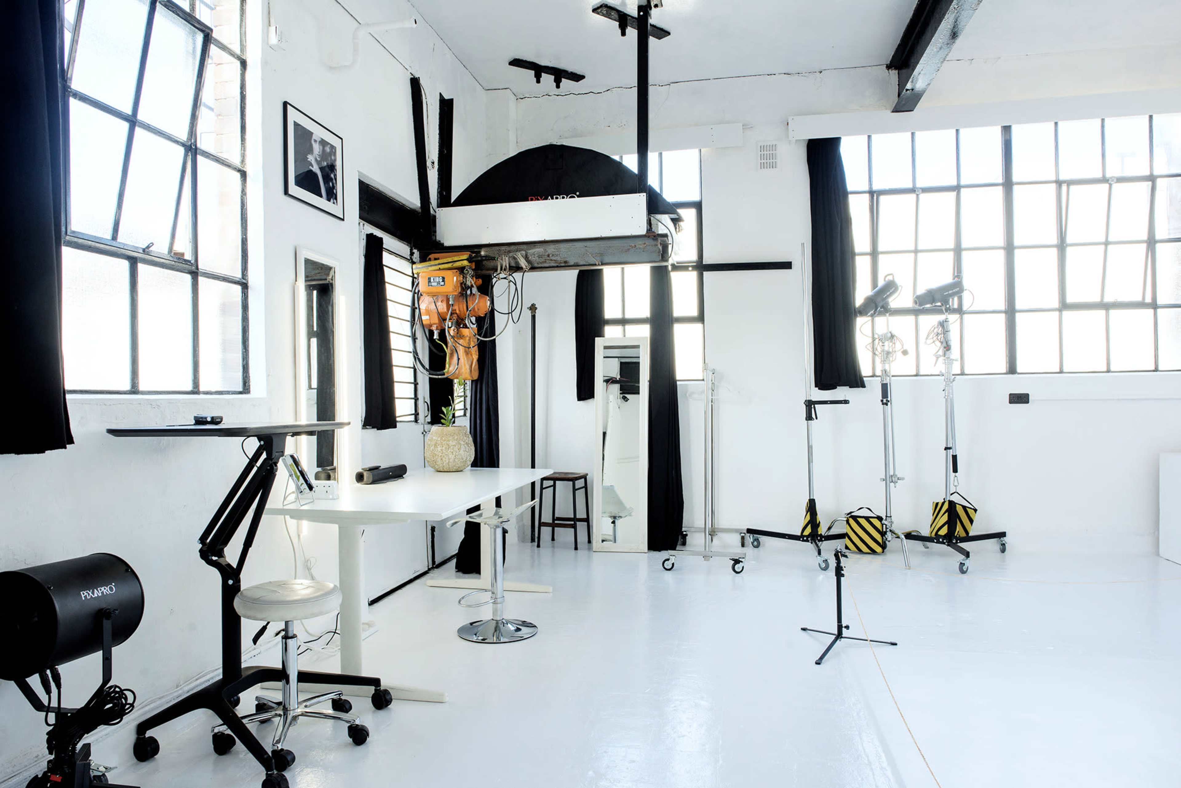 A bright, minimalist studio space with large windows, a table, a chair, and various lighting equipment.