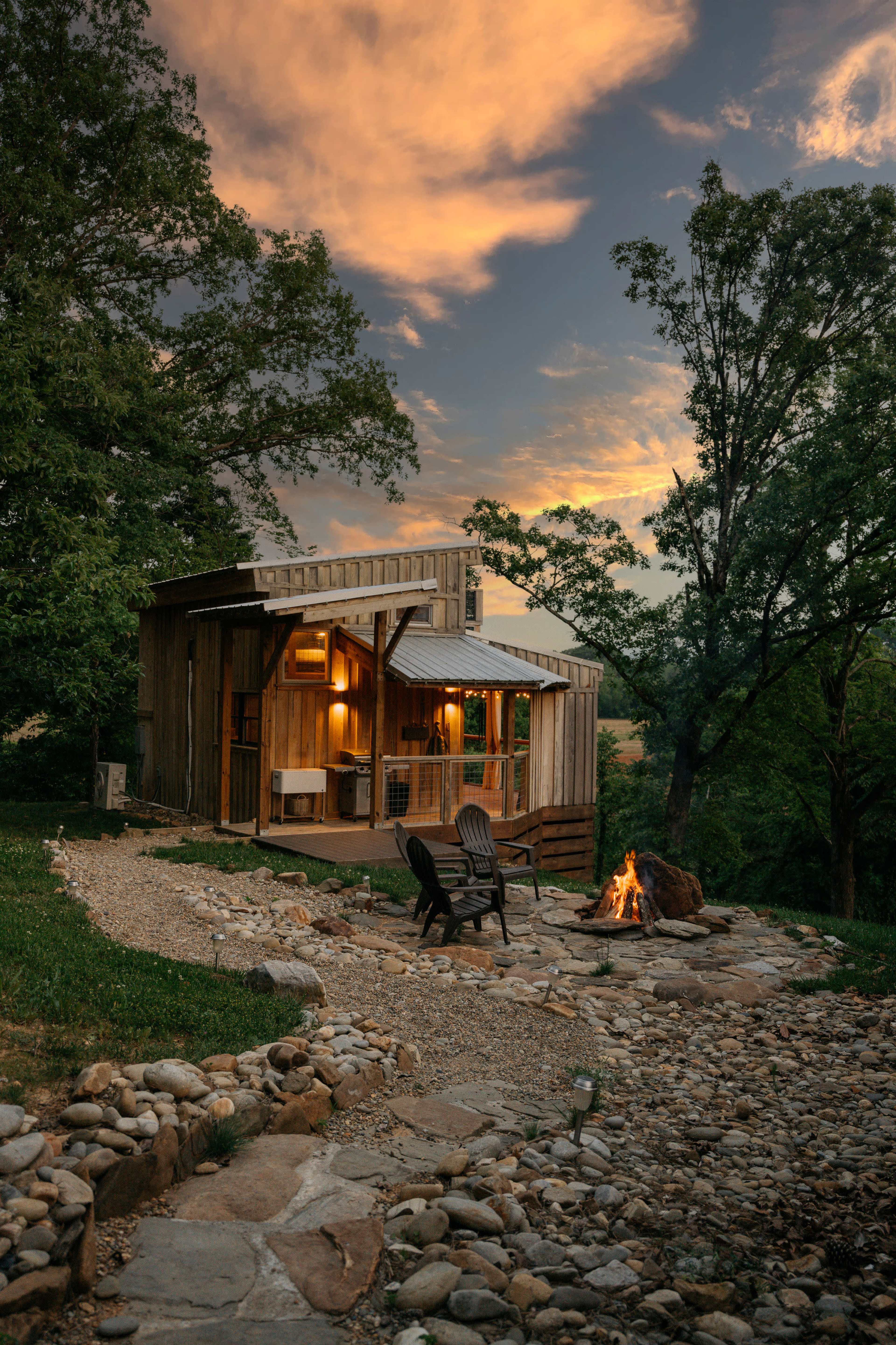 A wooden cabin with a stone path and fire pit is set against a sunset sky, surrounded by trees.