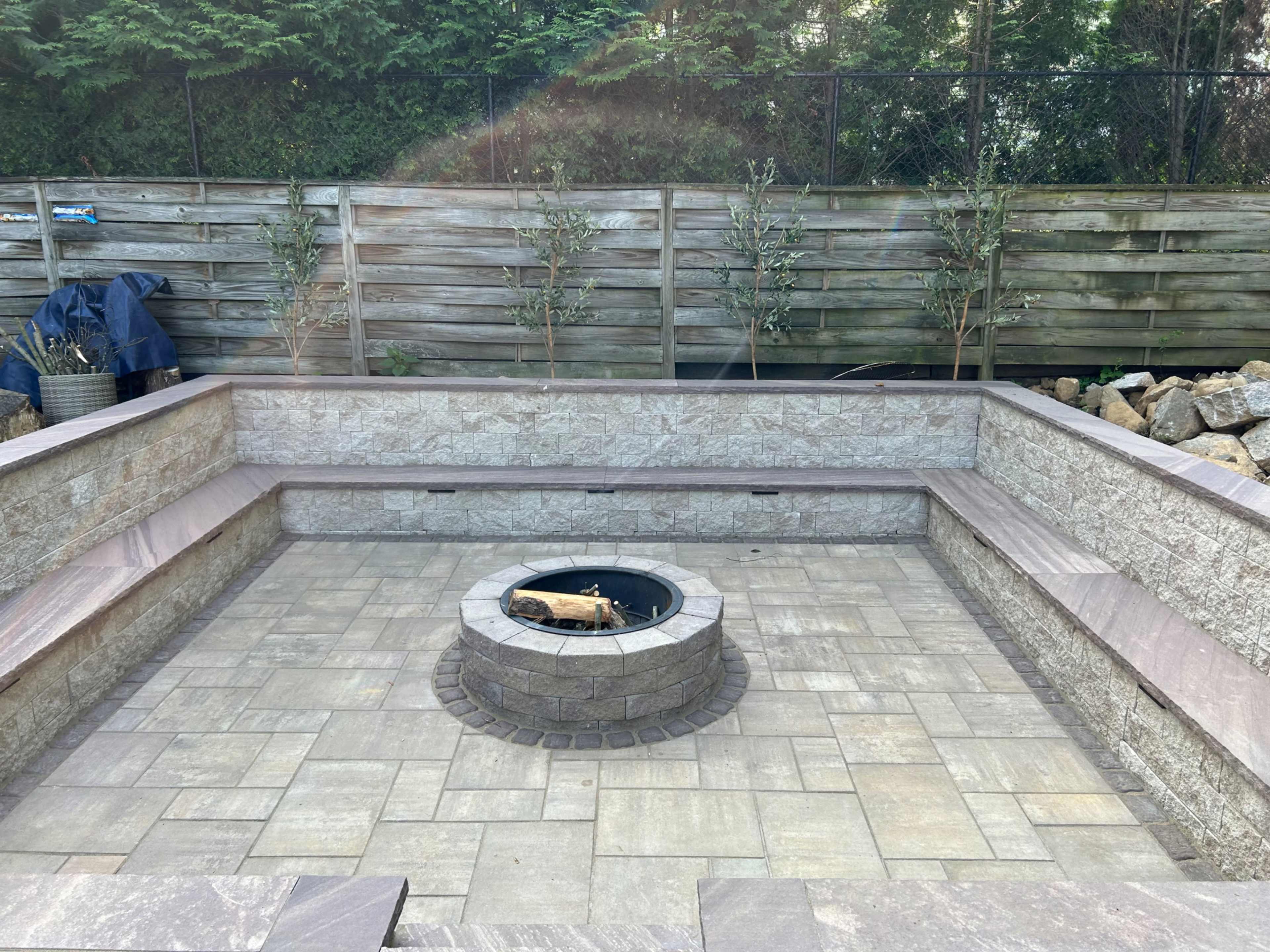 A circular stone fire pit surrounded by a square patio with low stone walls, set against a backdrop of wooden fencing and small plants.