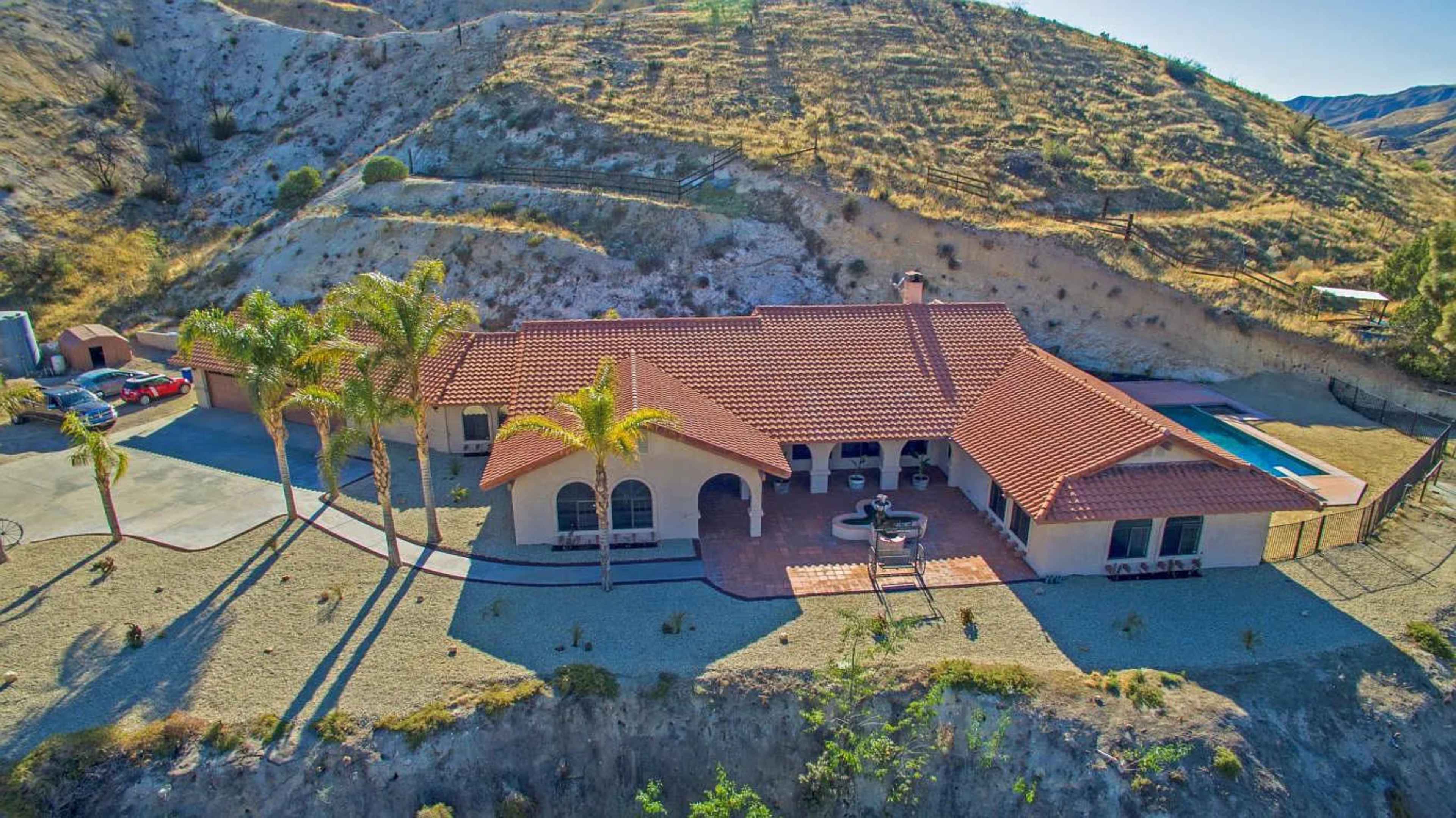 A single-story house with a red tile roof, situated on a hillside, surrounded by palm trees and a pool in the backyard.
