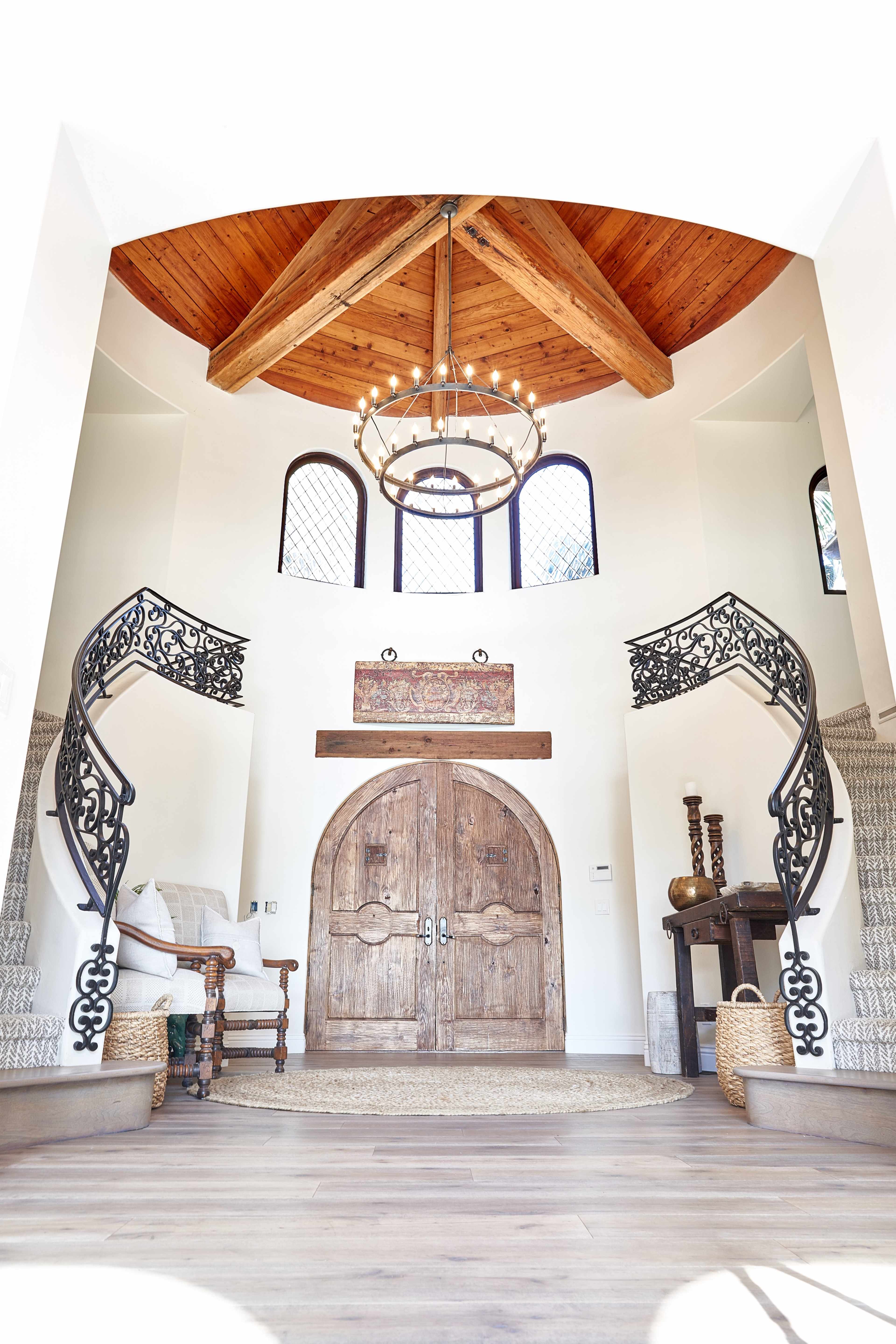 The image shows a spacious entryway featuring a high wooden ceiling, ornate iron railings on either side of a staircase, and a pair of large wooden doors.