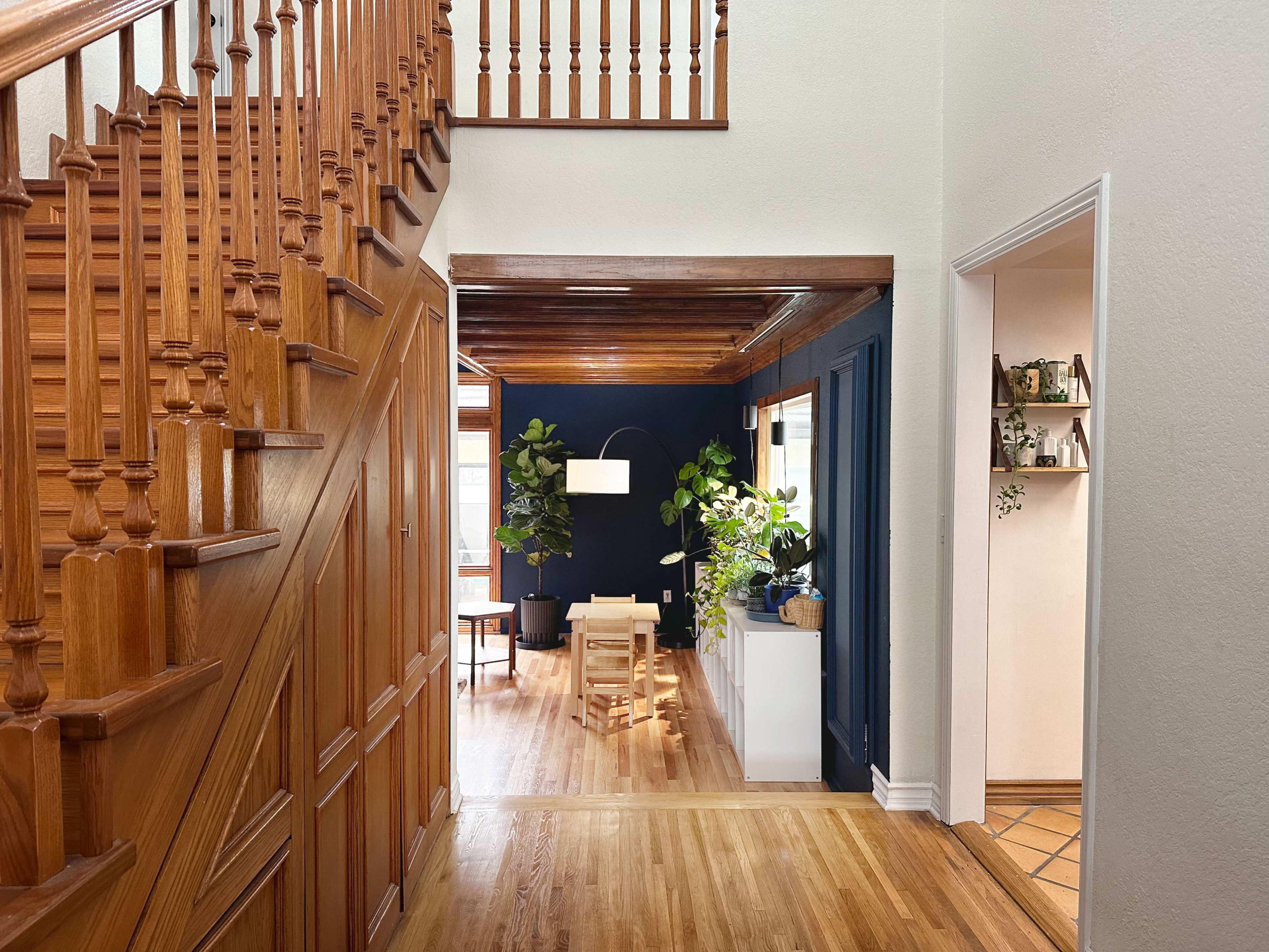 The image shows a wooden staircase leading to an open hallway that opens into a room with plants and a white lamp.