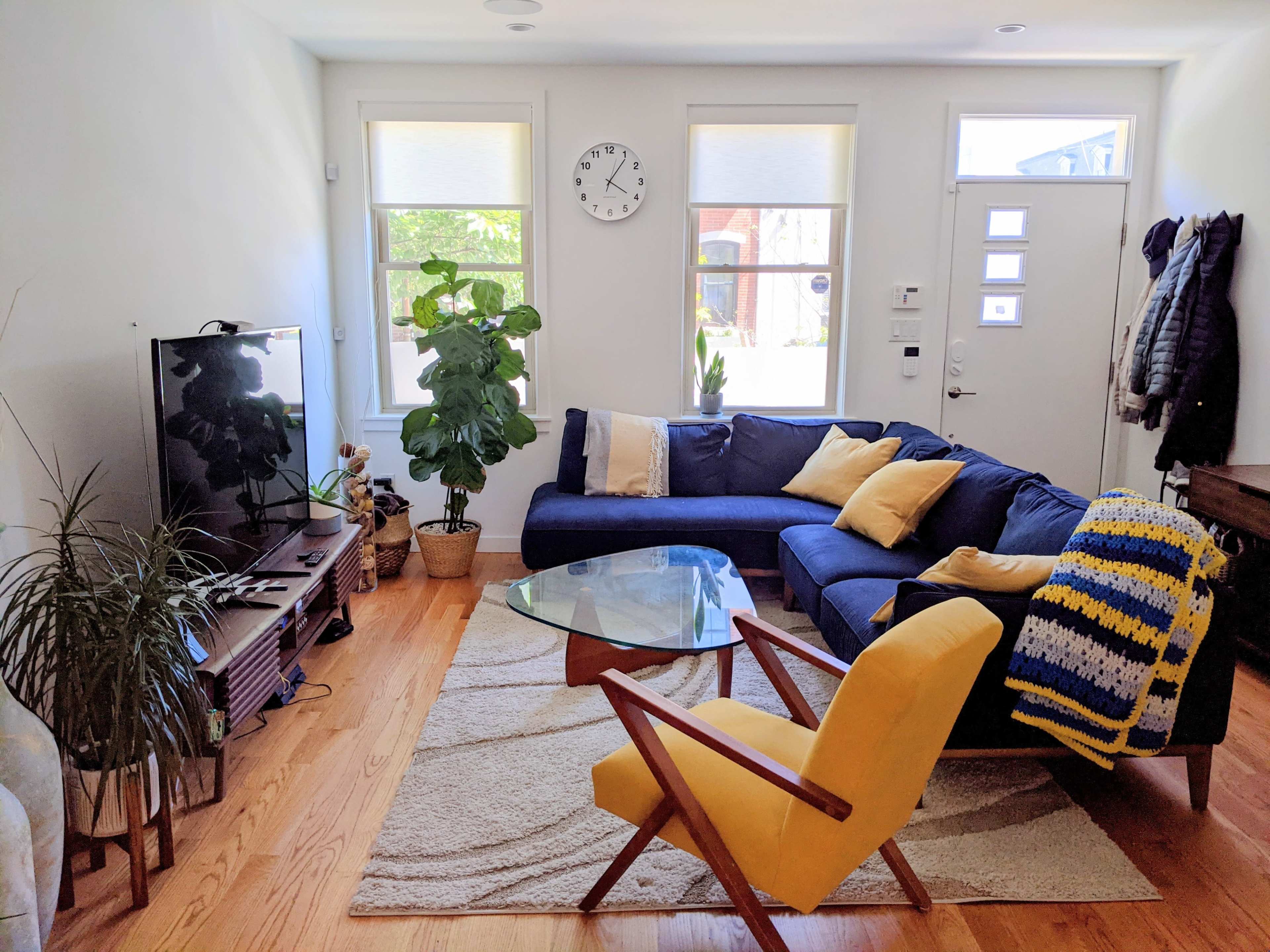 A living room featuring a blue sectional sofa, a glass coffee table, a television, and a yellow armchair, along with plants and a wall clock.