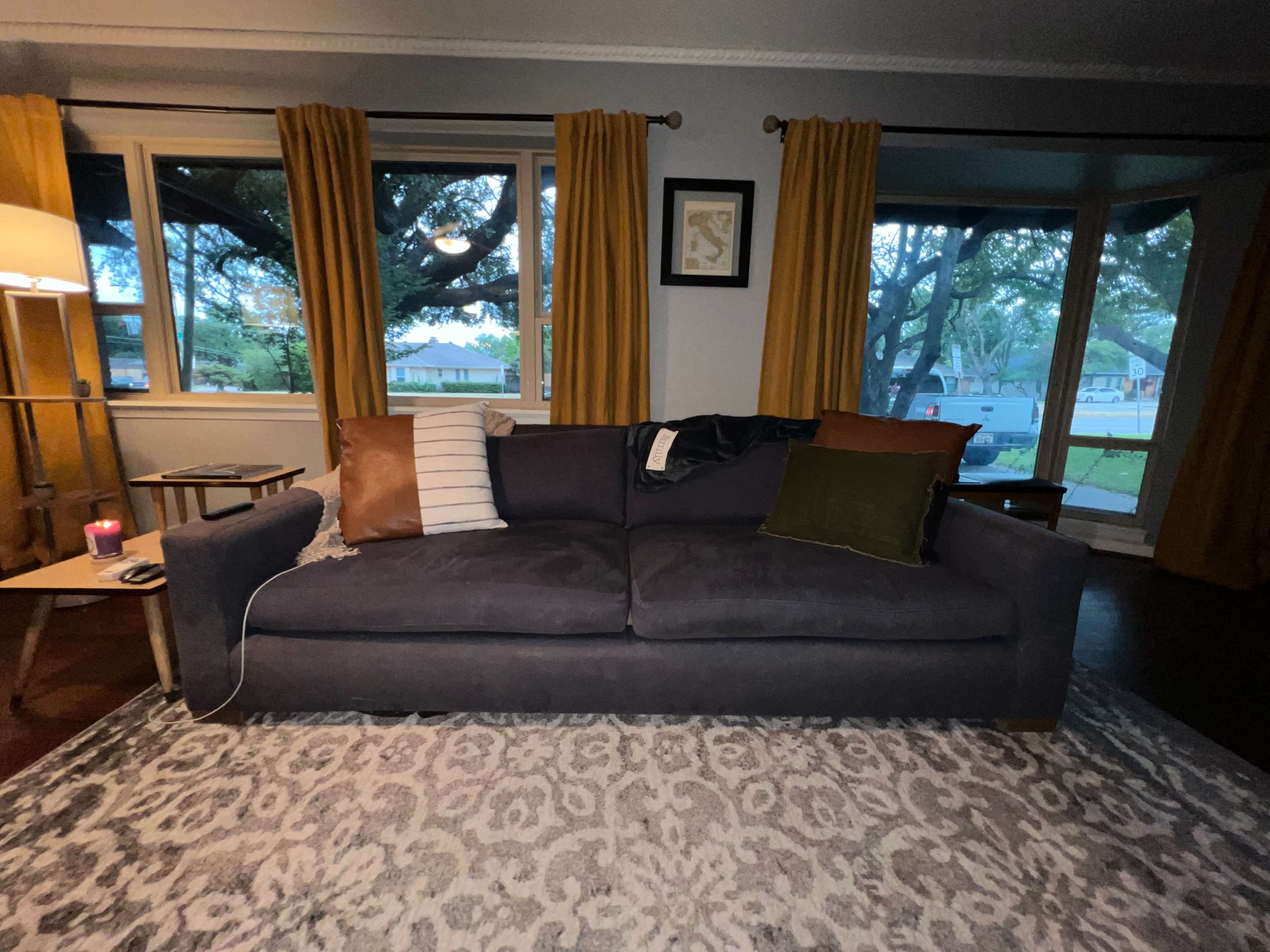 A dark sofa is positioned in a living room with yellow curtains and a patterned rug, near large windows that overlook a yard.