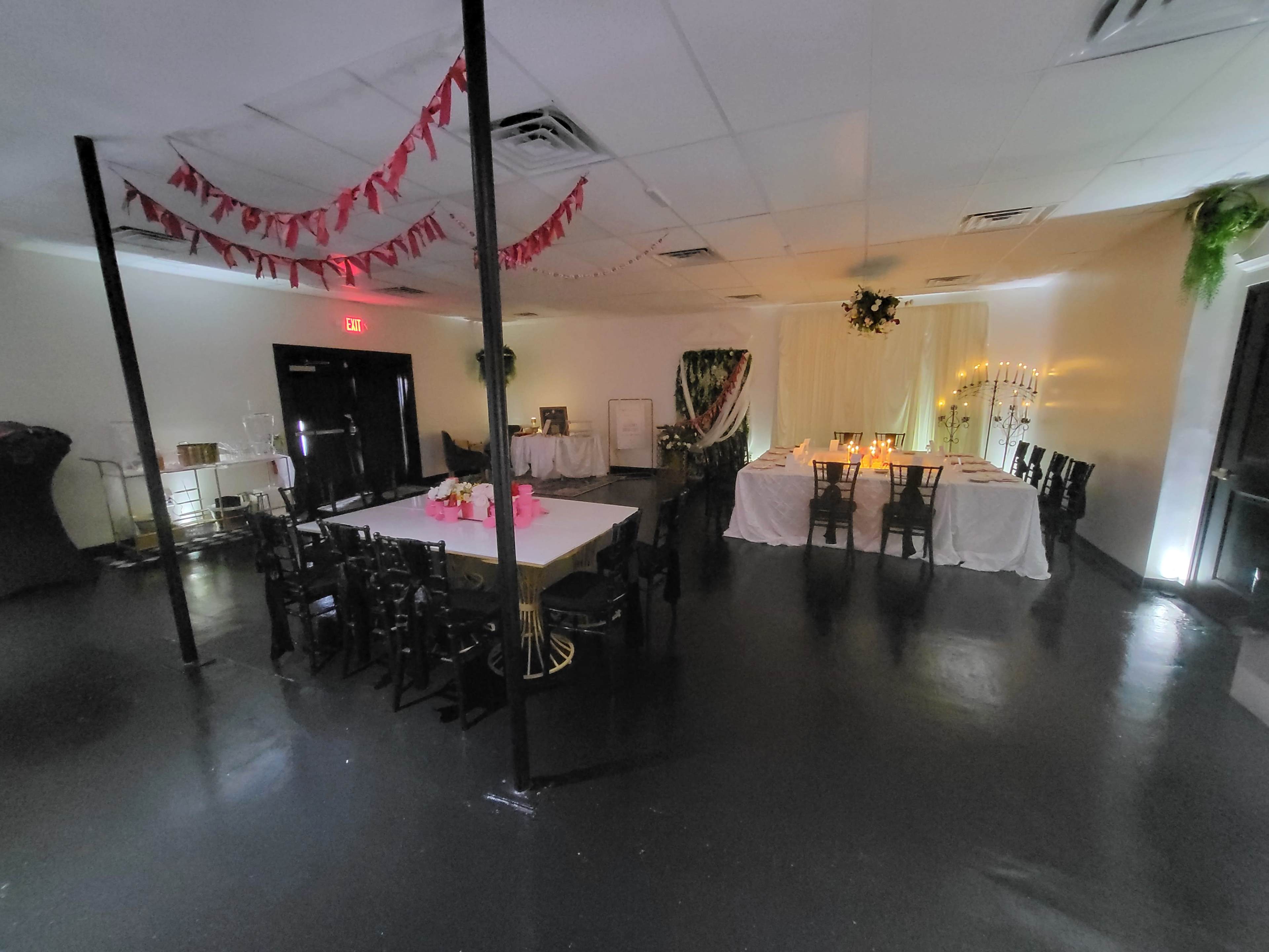 The image shows an indoor event space with tables and chairs set up, decorated with draped fabric and lighting, and a banner hanging from the ceiling.