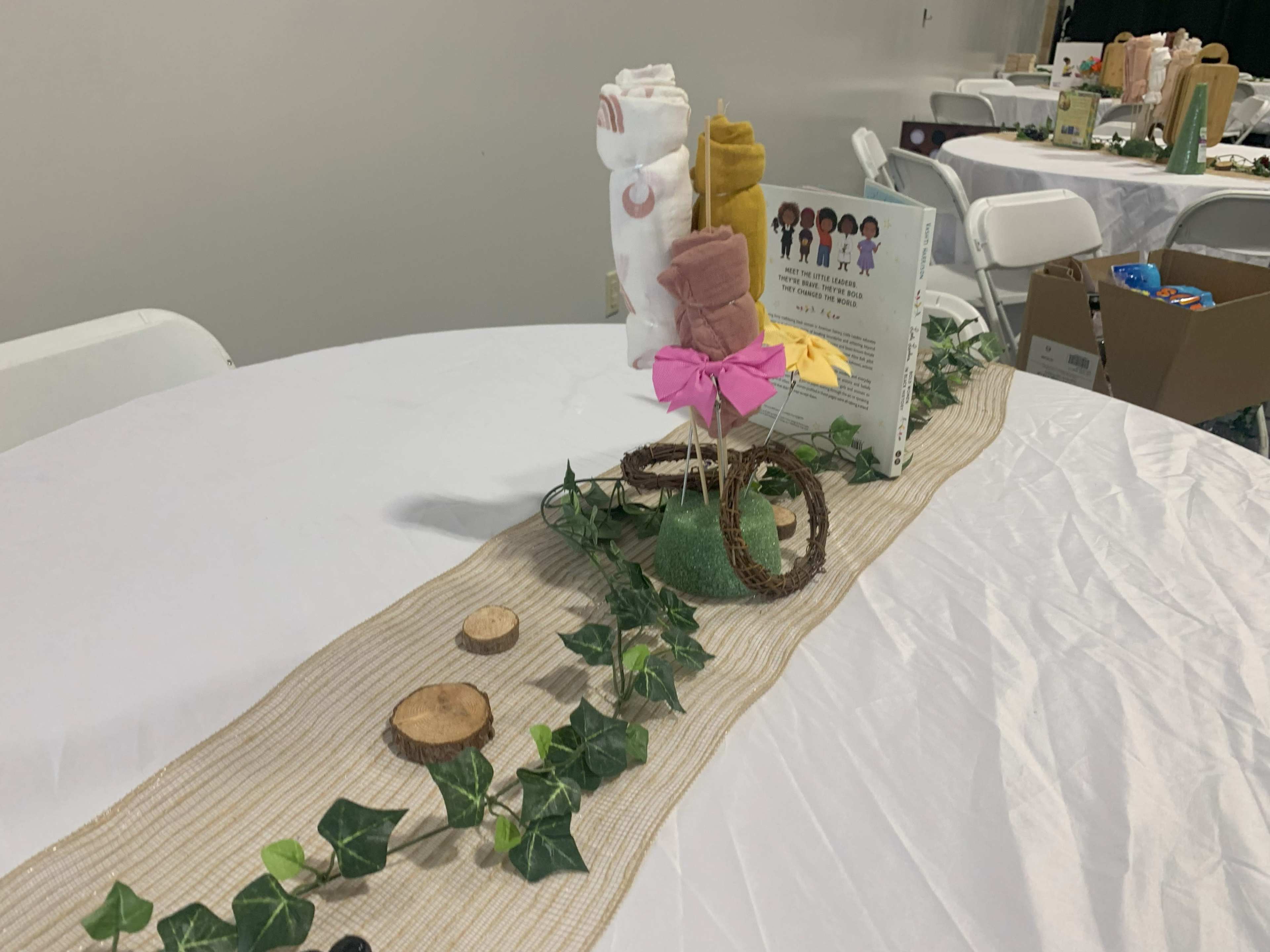 A table is set with a decorative centerpiece featuring rolled towels, a book, and a garland of artificial ivy.