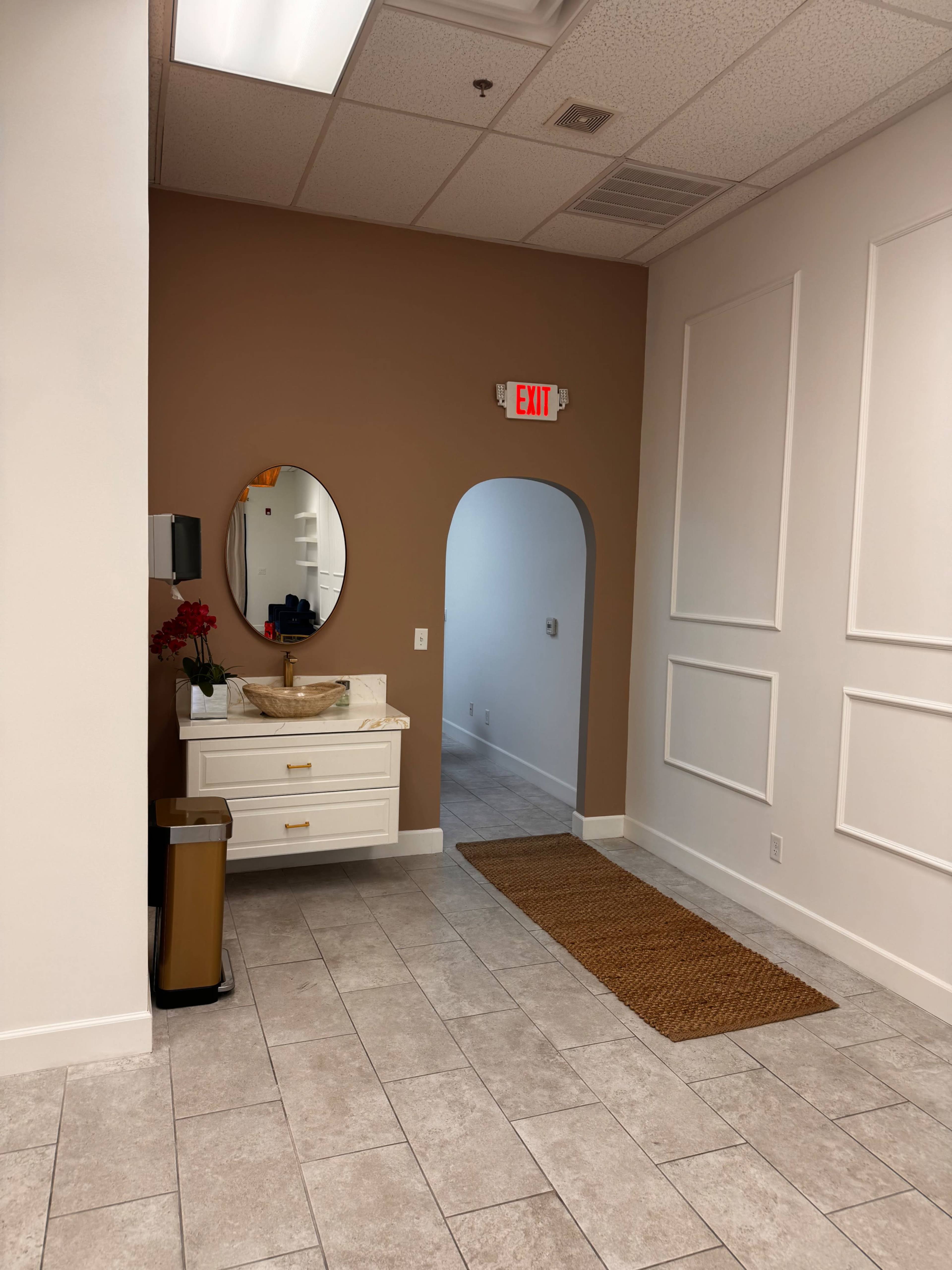 The image shows a well-lit room featuring a vanity with a round mirror, an exit sign, and a doorway leading to another area, with white and brown walls and tiled flooring.