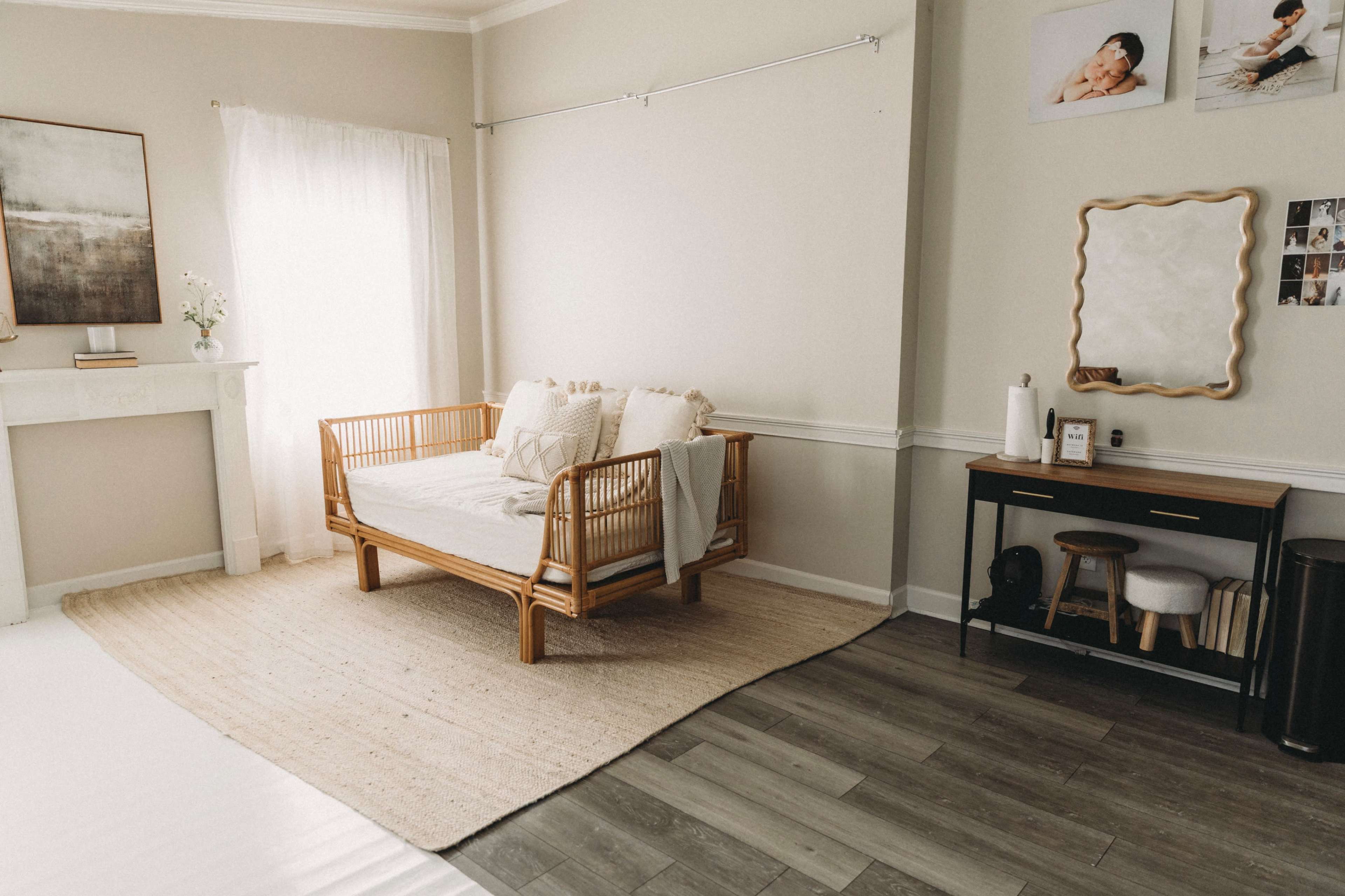 The image shows a light, airy room with a rattan daybed, a small wooden side table, and a rug on a hardwood floor.